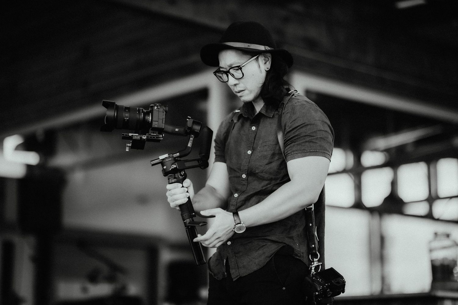 Black and white portrait of a wedding videographer in a hat and glasses operating a camera on a stabilizing gimbal.