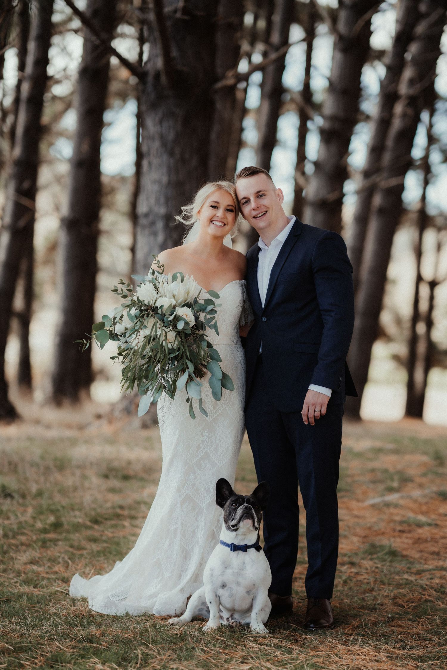 Smiling bride holding a green and white bouquet stands beside her groom in a navy suit with a French bulldog among pine trees.