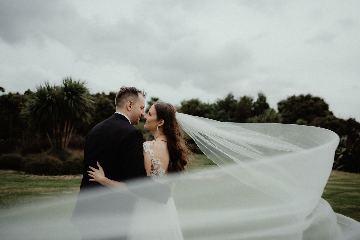 Groom and bride embrace on a grassy hillside as her sheer wedding veil billows dramatically in the wind under a moody sky.