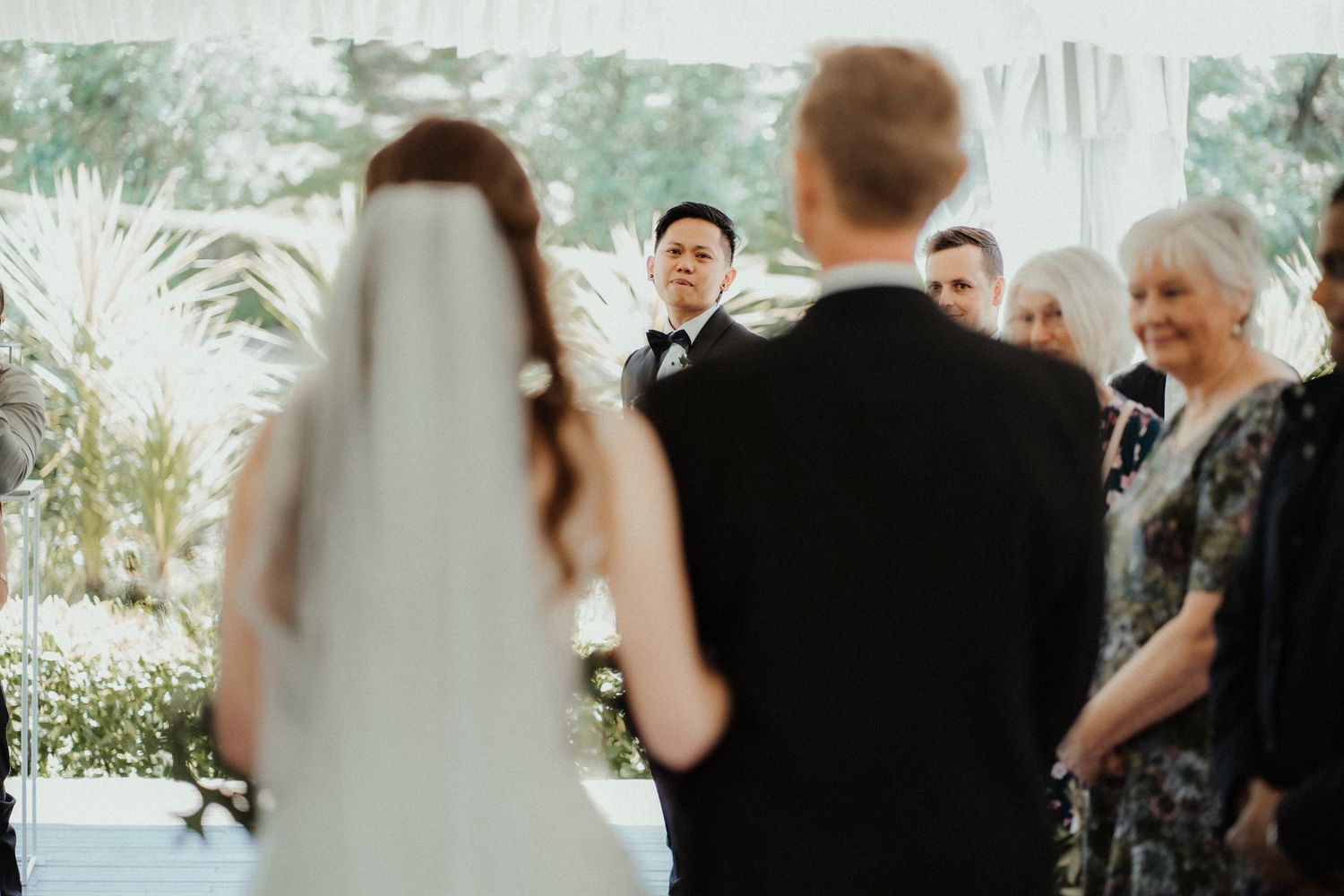 A bride in a long veil and groom stand facing the guests during an outdoor wedding ceremony as an officiant looks on.