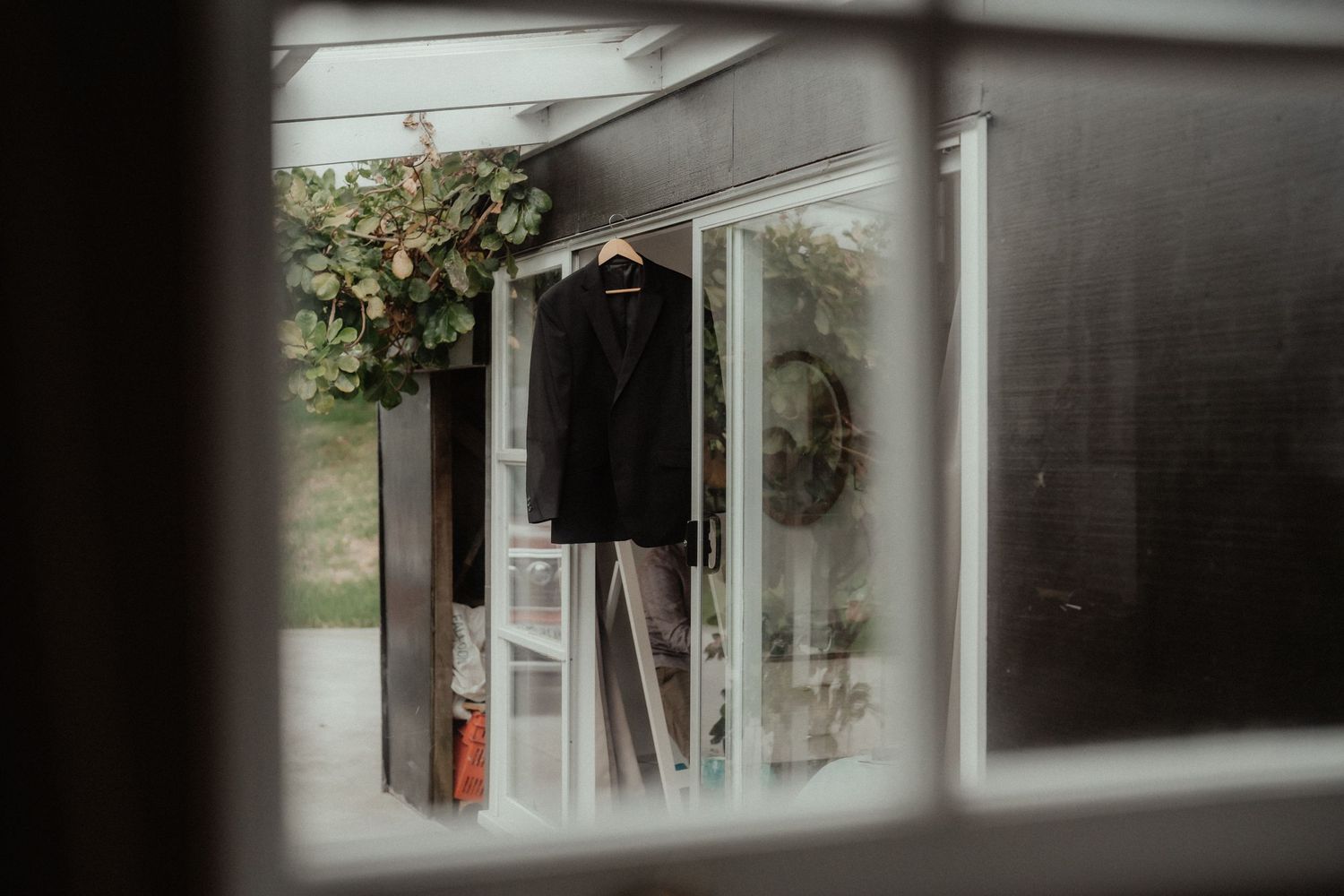 Groom's black suit jacket hangs on a rack, framed through a window with a floral wreath, captured before the wedding.