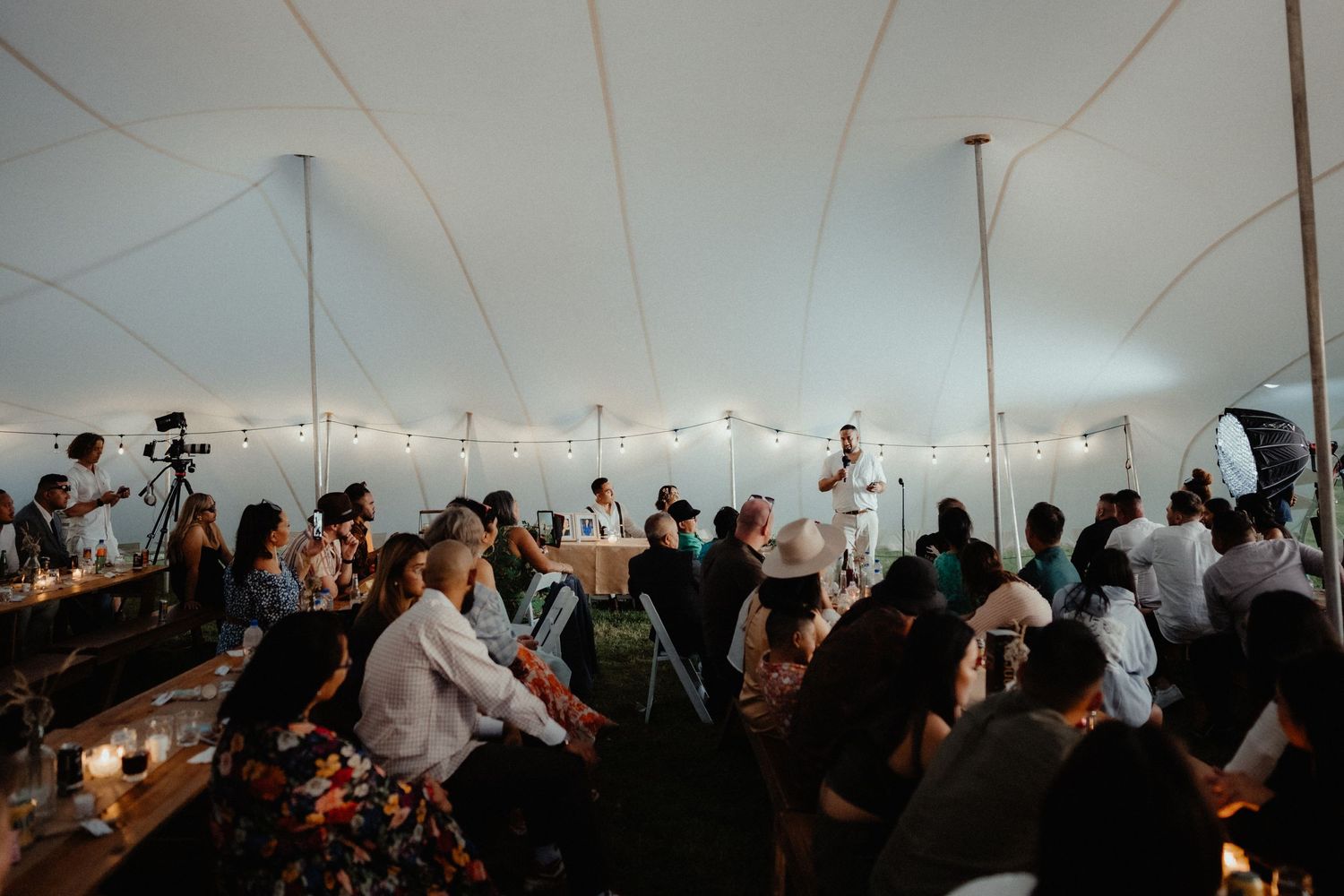 Guests sit at long tables under a white marquee tent with string lights while a speaker gives a toast at the wedding reception.