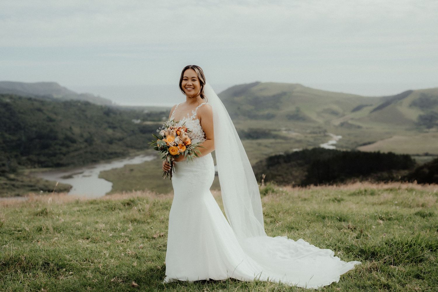 Smiling bride in a fitted white gown and veil holds an orange bouquet on a grassy hilltop overlooking a river and green valley.