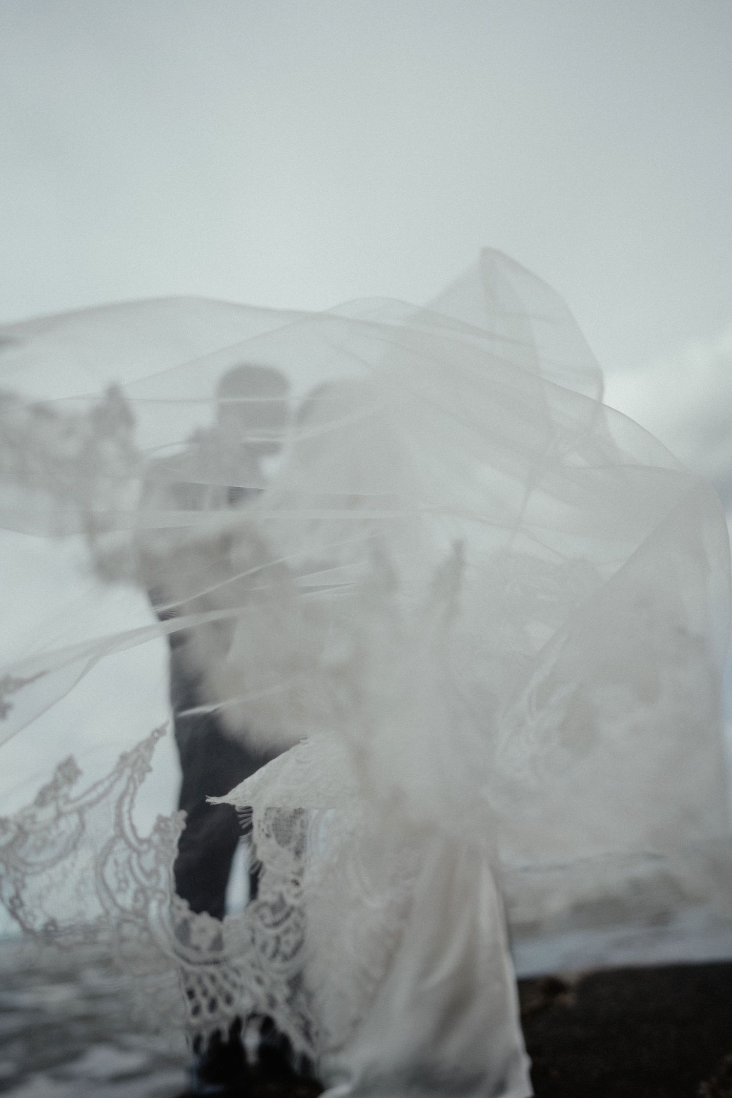 Wedding couple softly obscured behind a sheer lace-trimmed veil blowing in the wind under an overcast sky.