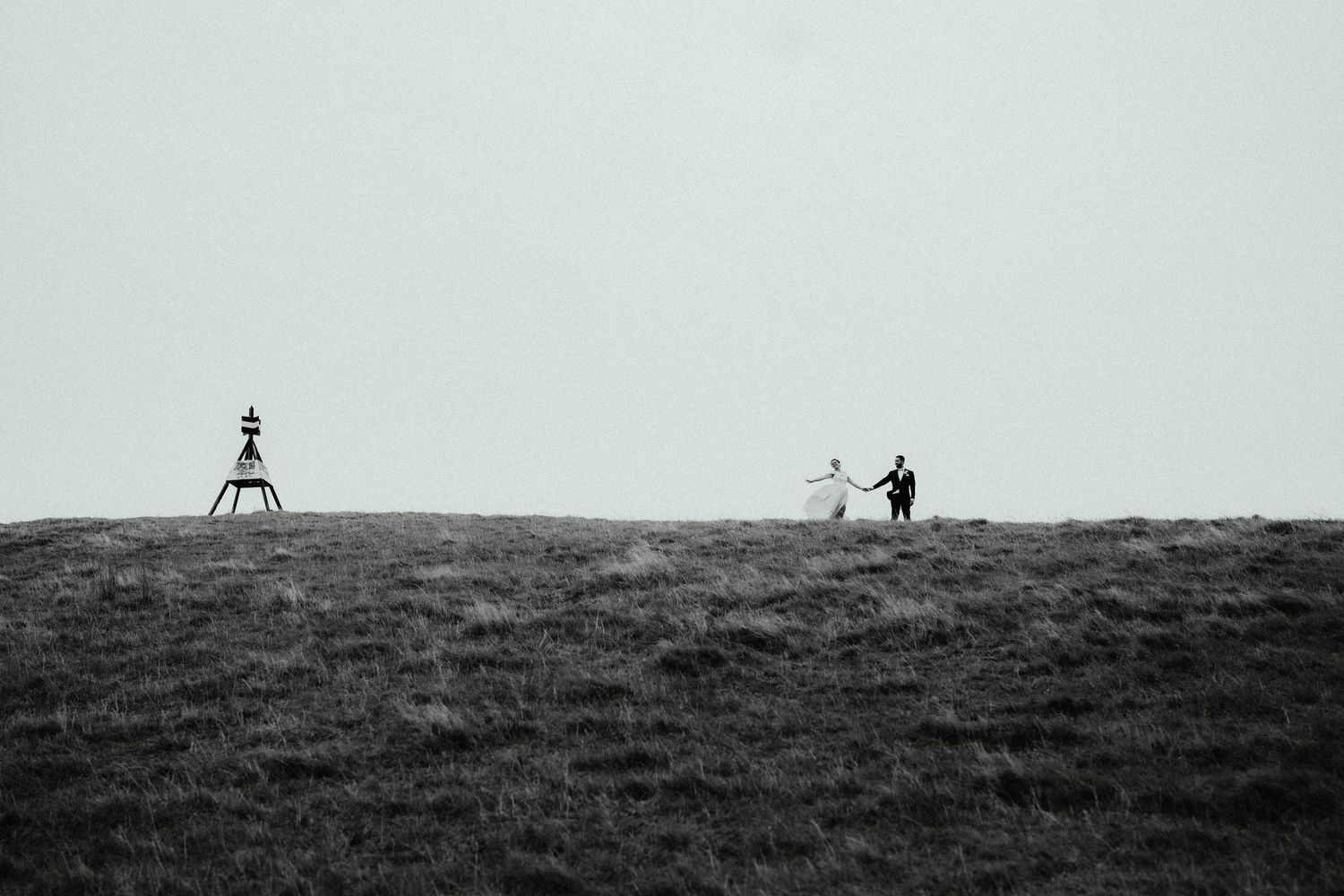 Tiny bride and groom hold hands on a grassy hilltop beside a trig beacon under a vast, minimalist black and white sky.