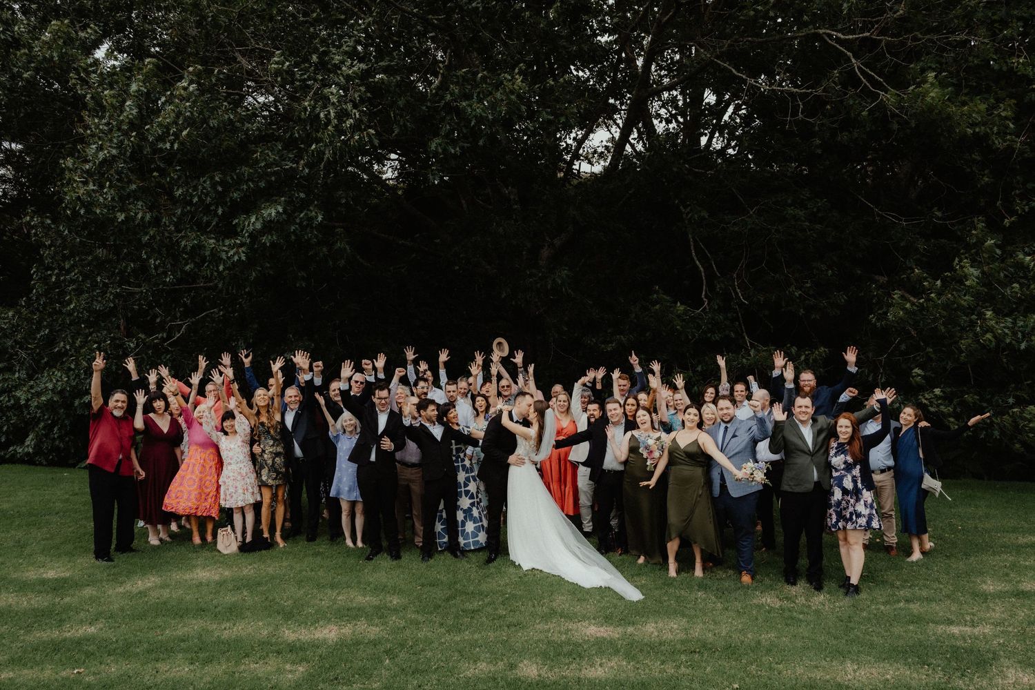 Bride, groom and cheering wedding guests pose for a joyful group photo on a green lawn in front of dark trees.