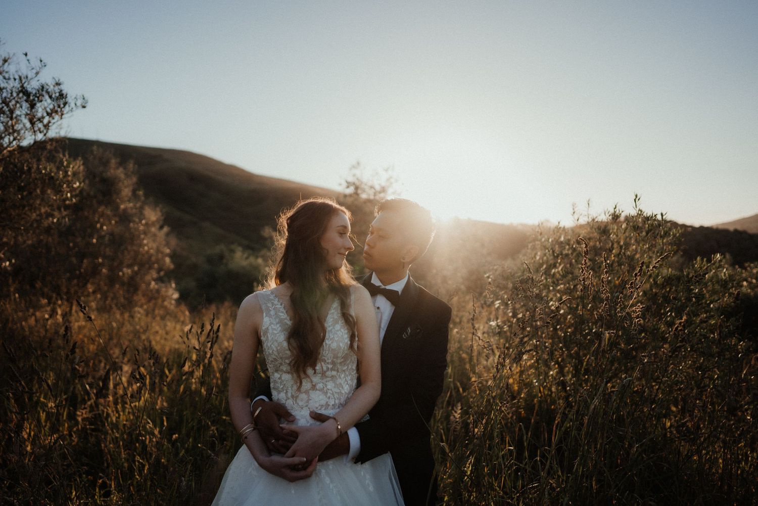 A bride and groom embrace in a hillside olive grove as the setting sun creates a warm, glowing backlight.