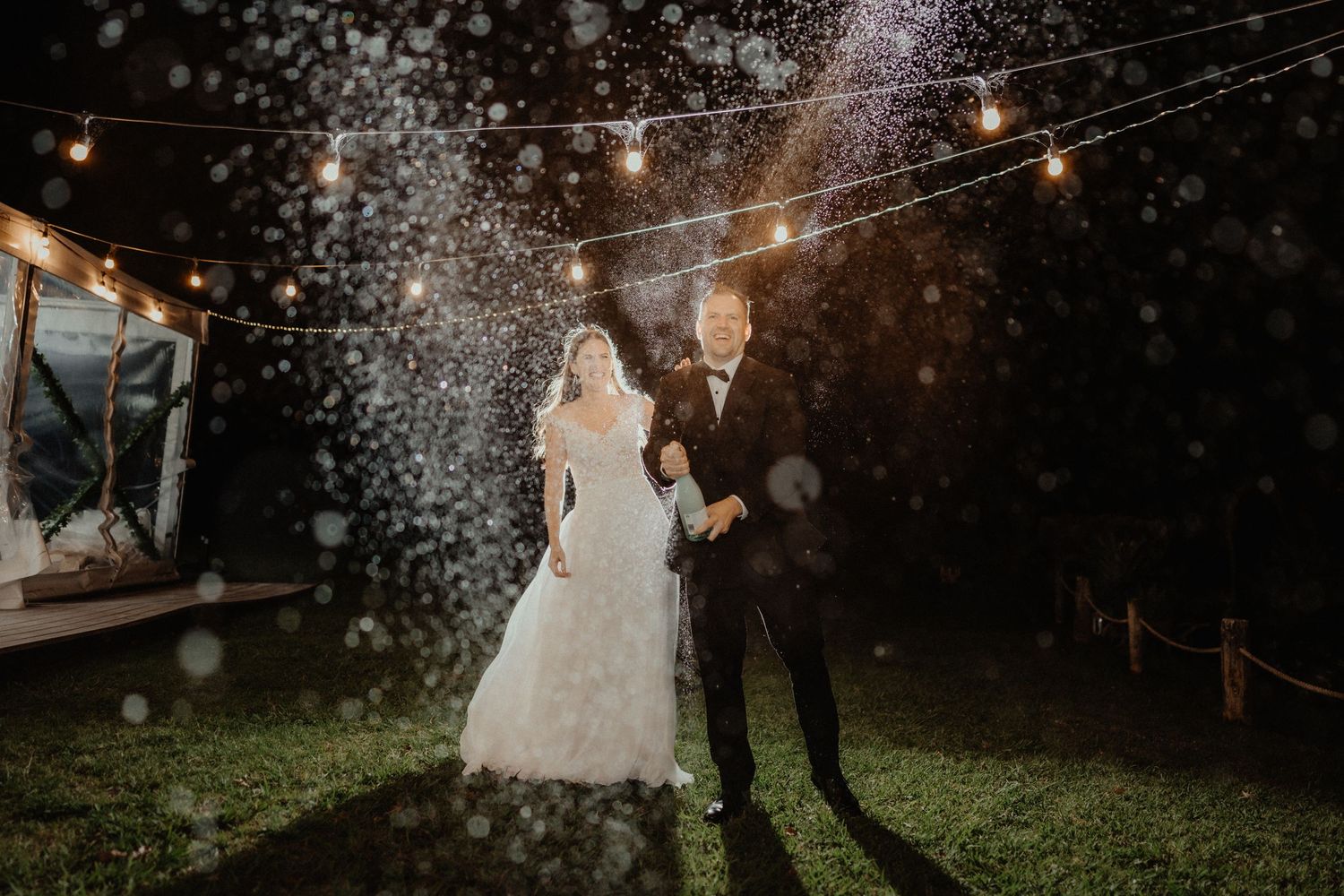 A bride and groom stand together on the grass at night as sparkling confetti fills the air beneath strings of warm festoon lights.