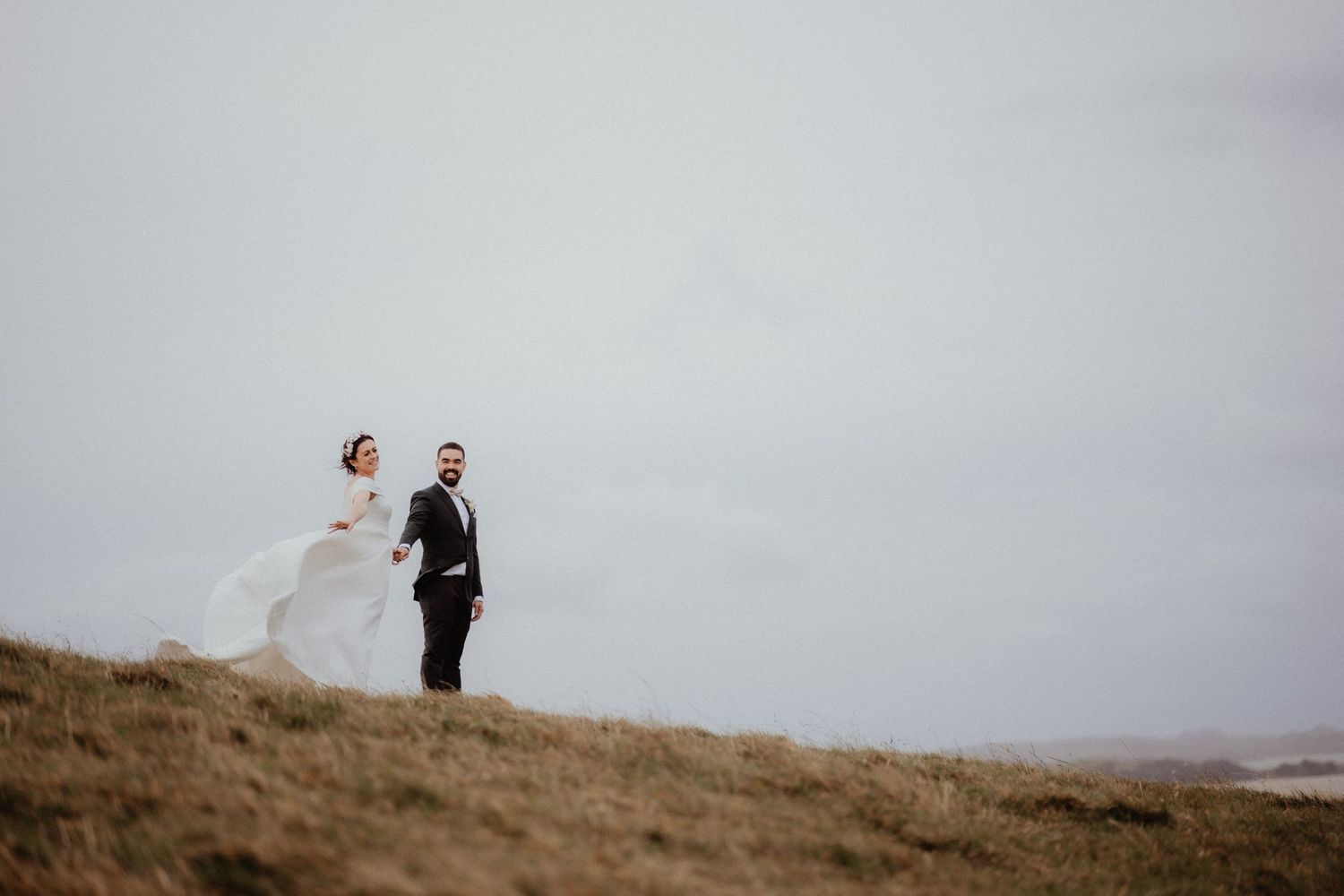 Bride and groom hold hands while walking along a windswept grassy hilltop under a soft gray sky.