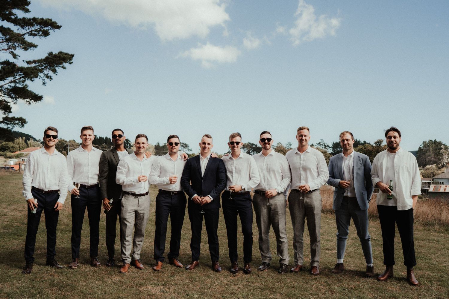 Groom stands with eight groomsmen in a line on a grassy hillside under a blue sky, dressed in white shirts and dark trousers.