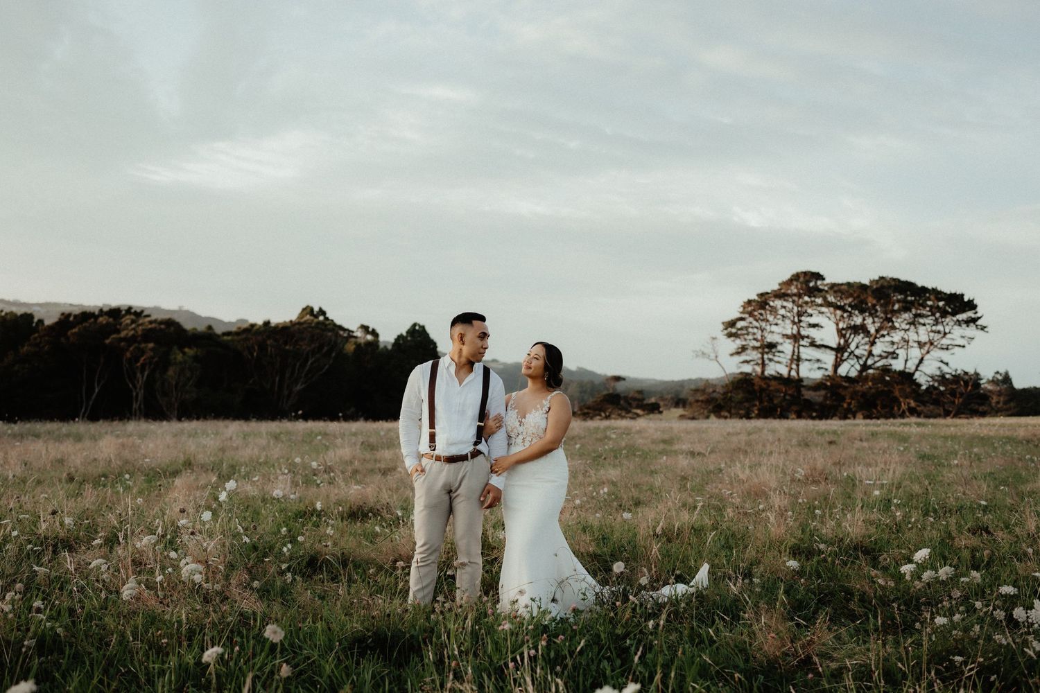 Bride and groom embrace in a wide meadow at dusk with trees and a soft cloudy sky behind them.
