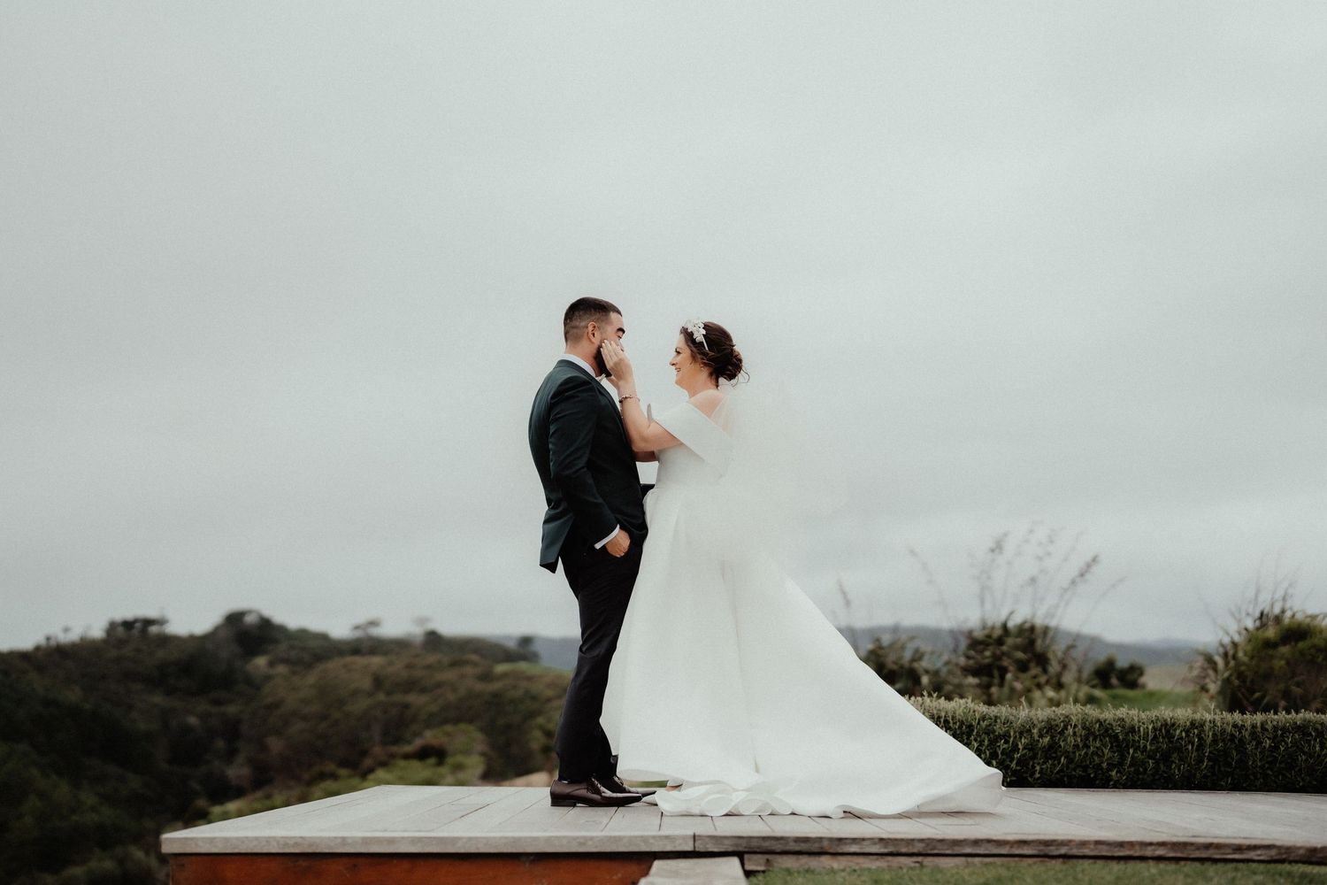 Bride in a white gown with a long veil and groom in a dark suit share a tender moment on a wooden platform with hilltop views.