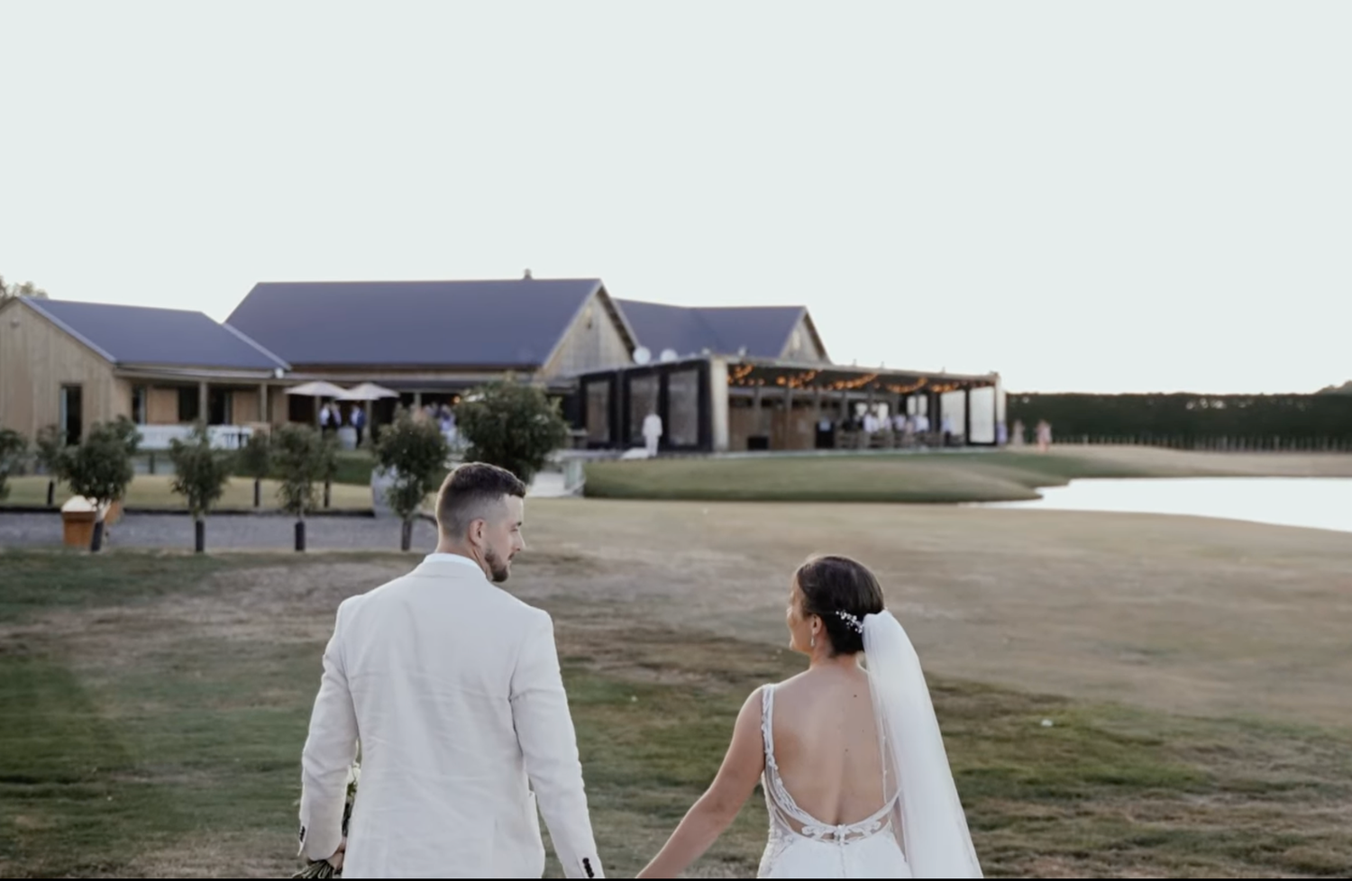 Newlyweds walk hand in hand across a lawn toward a modern countryside wedding venue beside a calm lake at dusk.