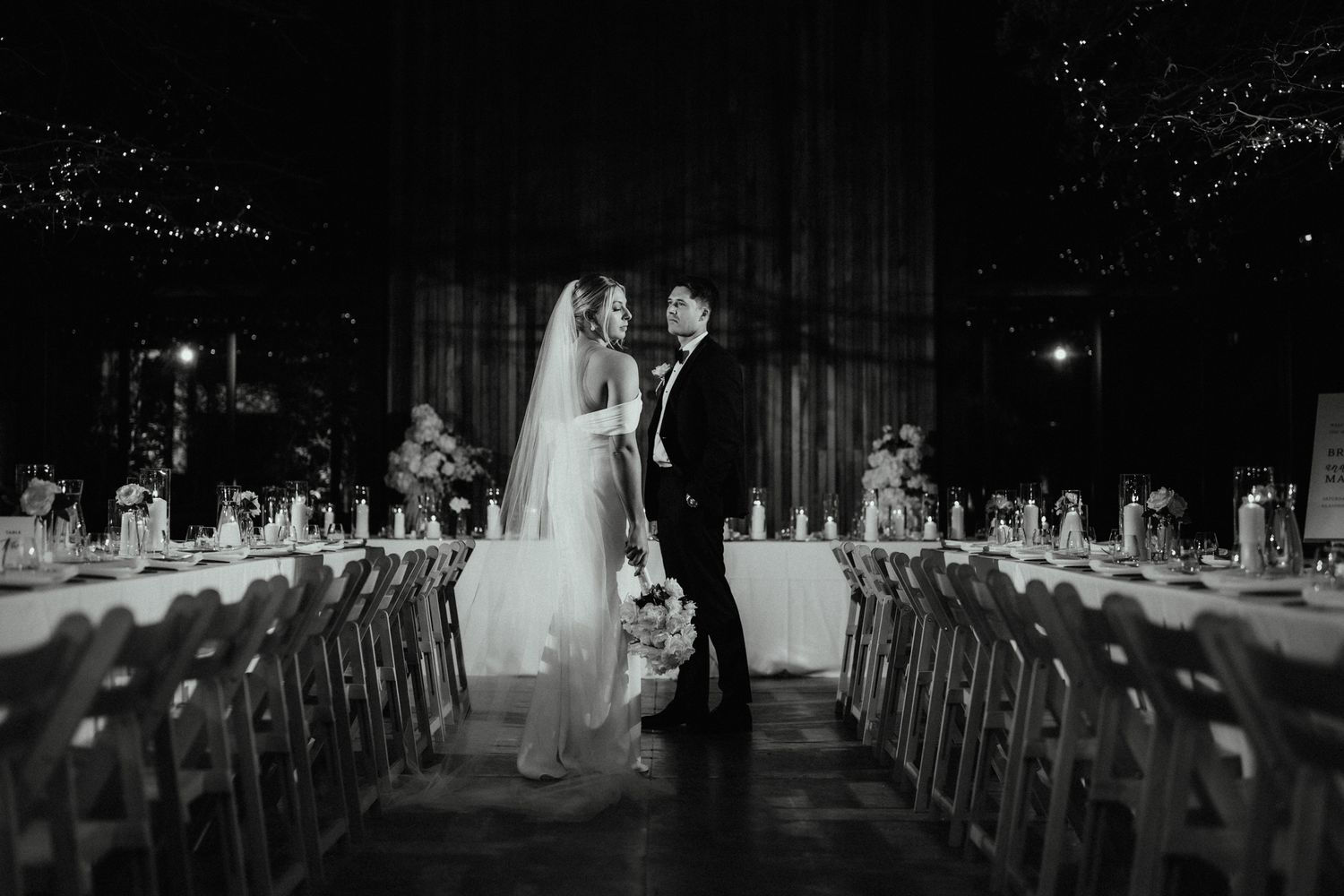 Black and white photo of a bride and groom standing together in a dimly lit reception hall with long tables and string lights.