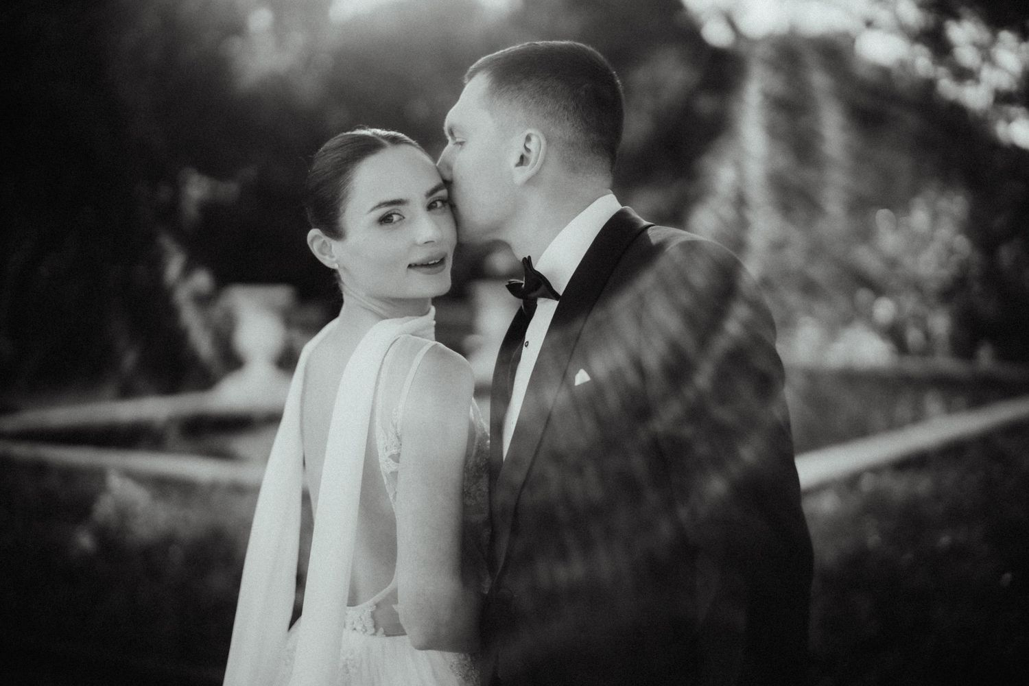 Black and white wedding photo of a groom kissing the cheek of his bride, who looks over her shoulder in a garden setting.