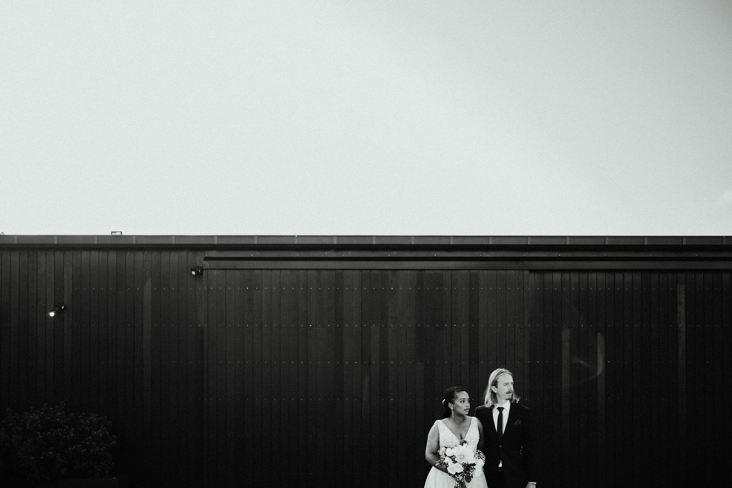Black and white wedding portrait of a bride and groom standing side by side against a dark wooden wall under a wide open sky.