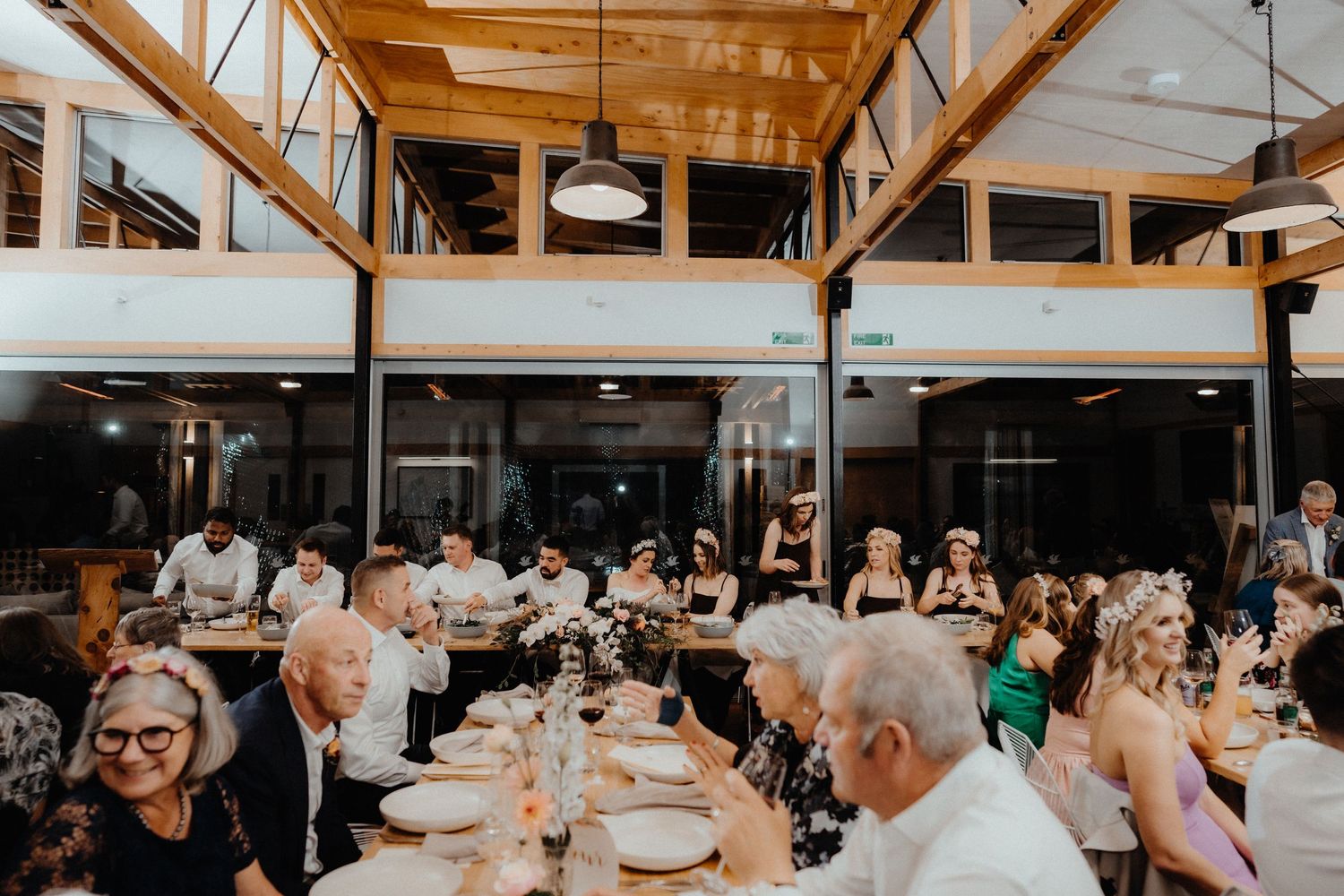 Wedding guests dine at long tables under wooden beams in a bright reception venue with floral centerpieces.