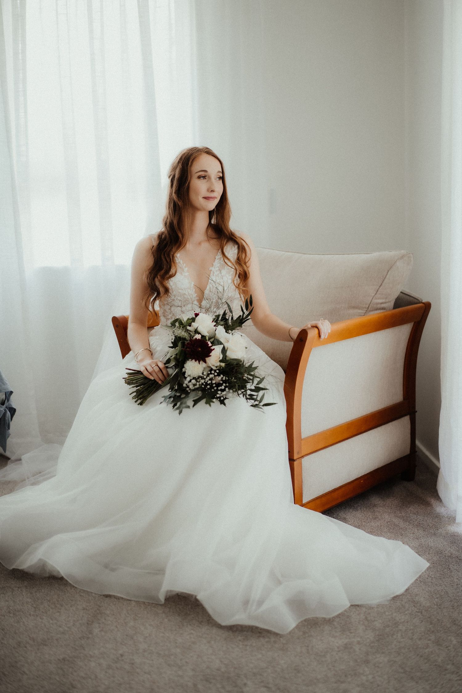 Bride in a lace and tulle wedding gown sits in a wooden chair holding a white and burgundy bouquet beside sheer curtains.
