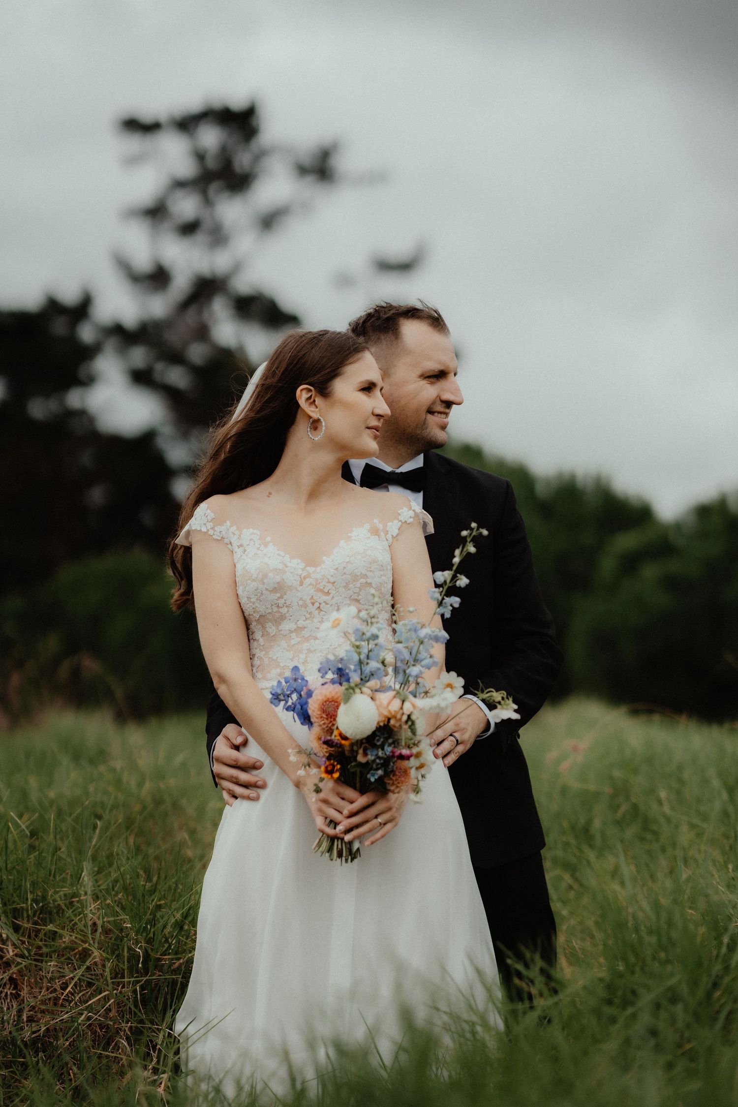 Groom embraces his bride from behind in a grassy meadow as she holds a colorful wildflower bouquet.