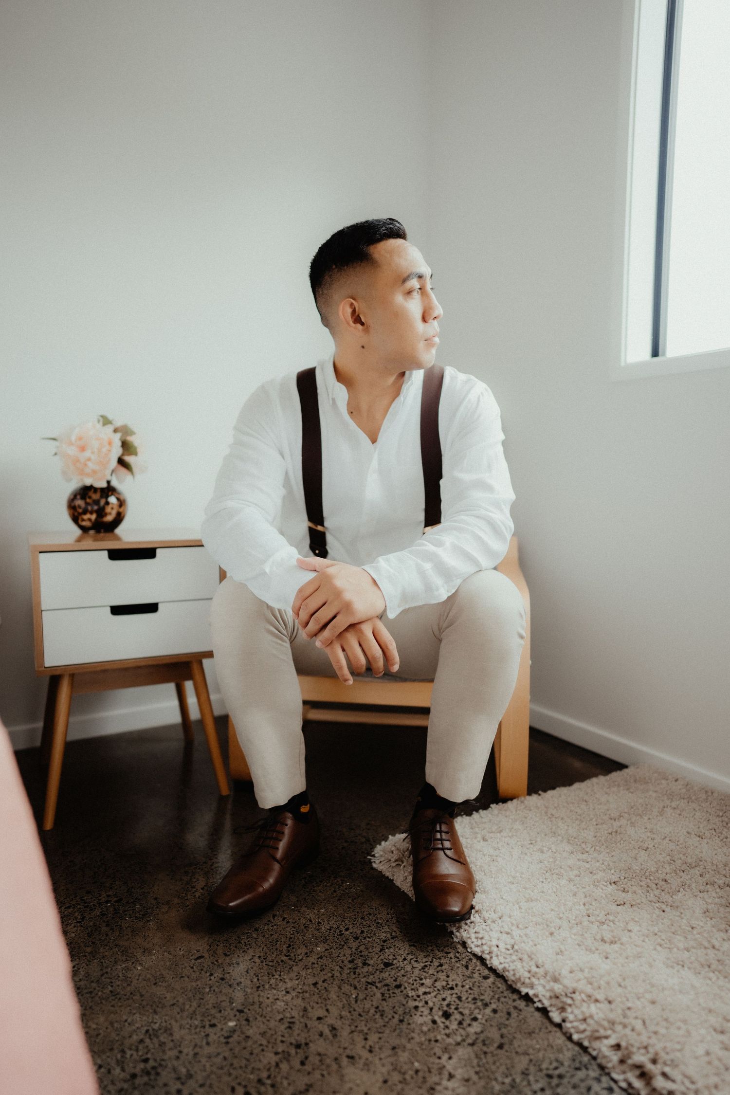 Groom in a white shirt, suspenders and brown leather shoes sits on a wooden chair in a bright room, gazing out the window.