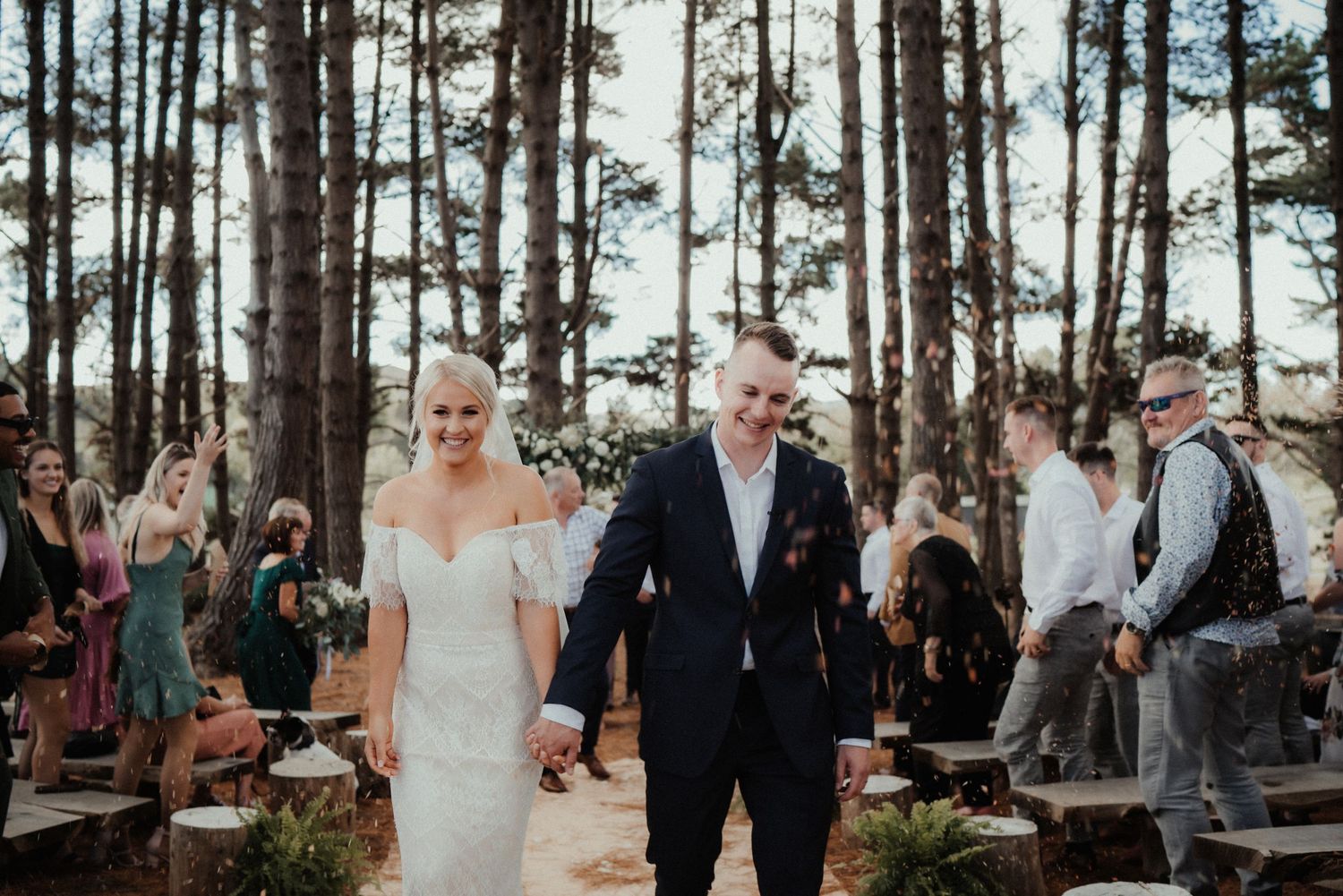 Newlyweds walk hand in hand down the aisle as guests cheer and smile among tall pine trees after their ceremony.