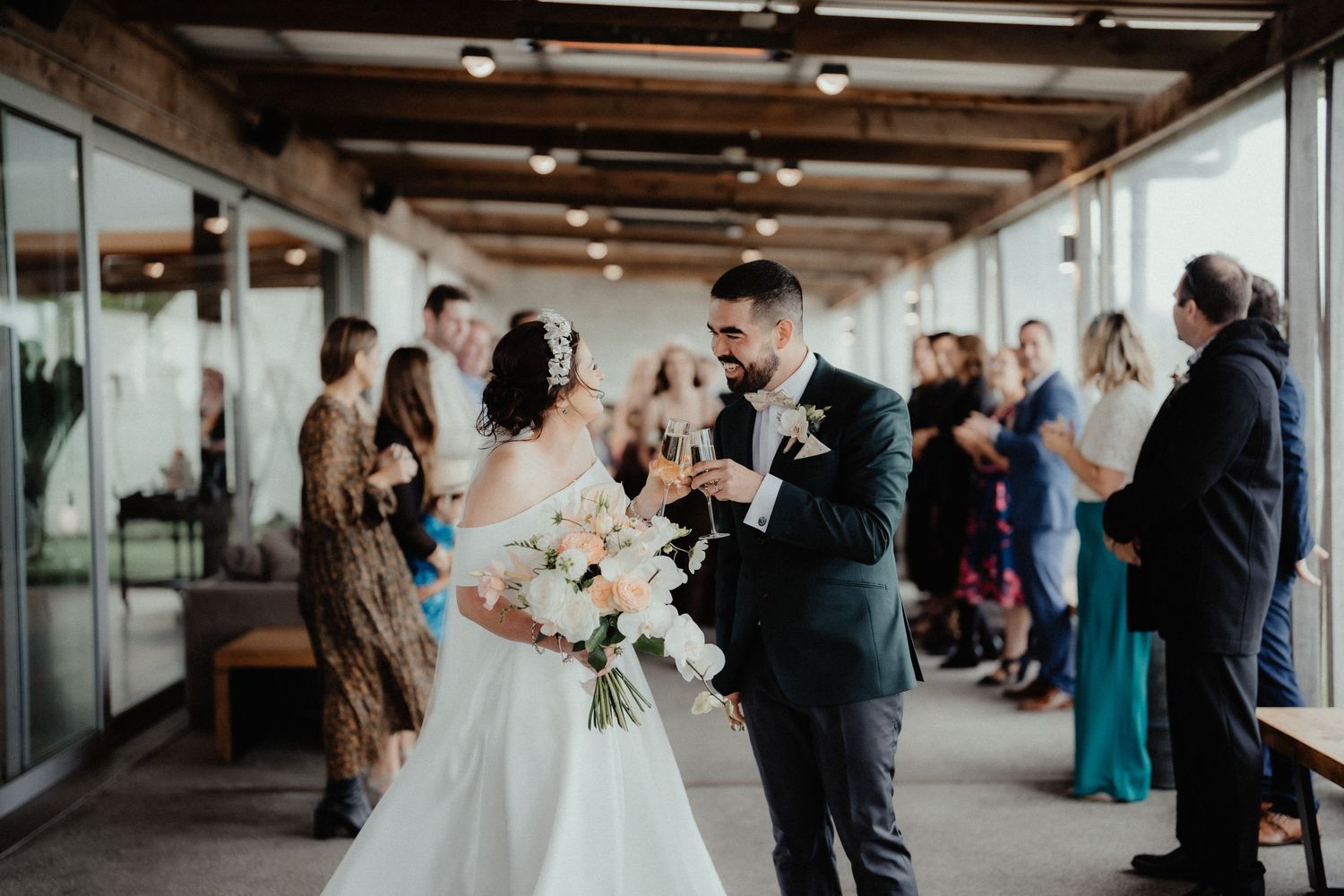 Smiling bride and groom walk down the aisle holding a bouquet and champagne glasses as wedding guests cheer in a bright venue.
