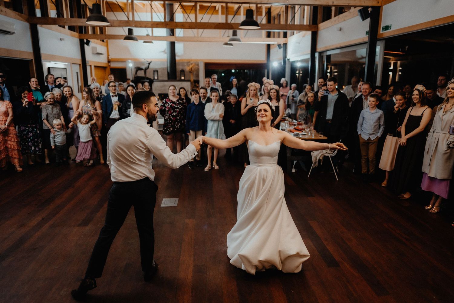 Bride and groom dance their first dance on a wooden floor while smiling wedding guests watch from around the room.