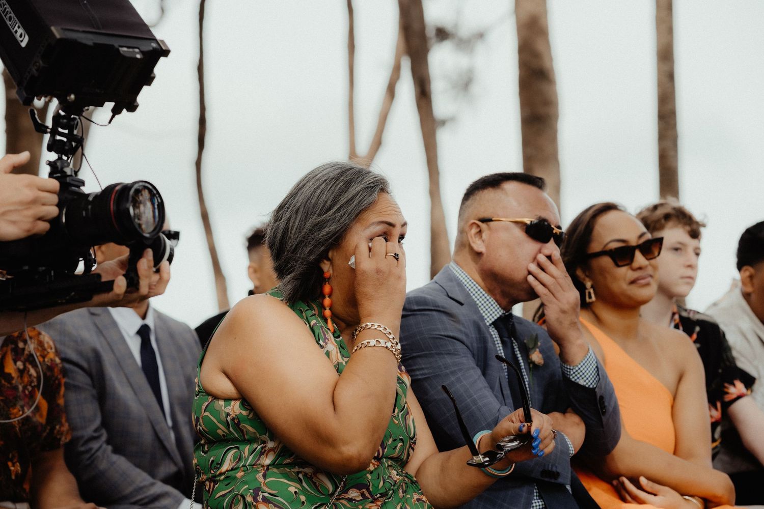 Emotional wedding guests wipe away tears during the ceremony while a videographer films from the left.