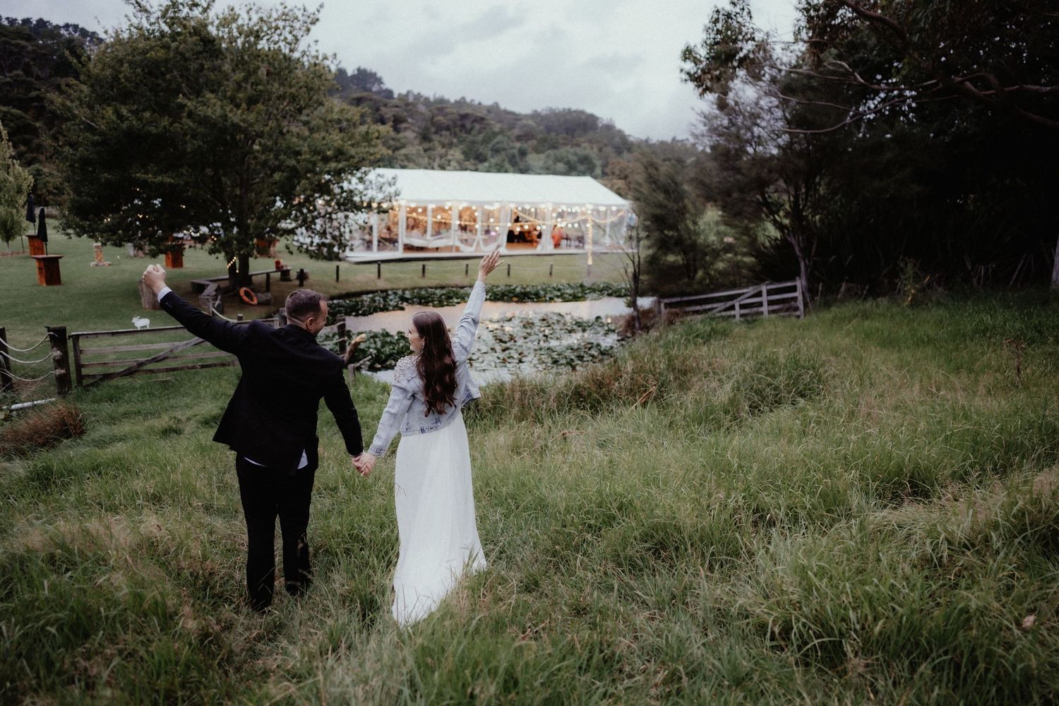 Bride and groom hold hands and raise their arms while walking through a grassy field toward a white wedding marquee.