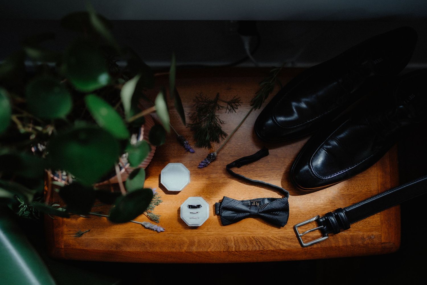 Groom's black leather shoes, bow tie, belt and ring boxes arranged on a wooden table beside green foliage.