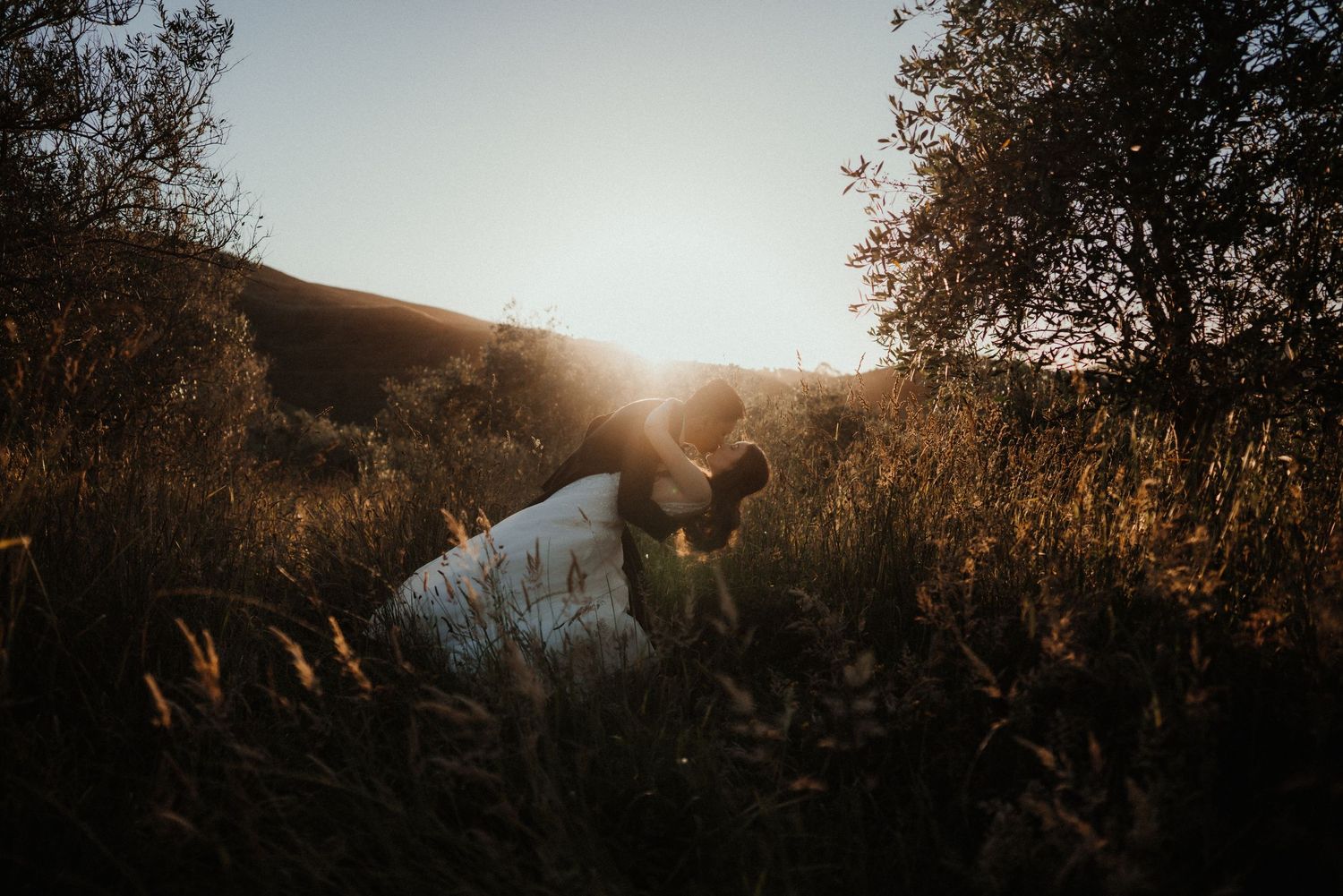 Couple in wedding attire kneel in tall golden grass as the sun sets behind a hill, creating a warm glow.