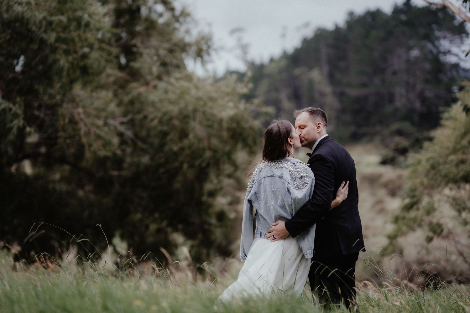 A bride and groom embrace and kiss in a grassy field with trees behind them during their outdoor wedding photoshoot.