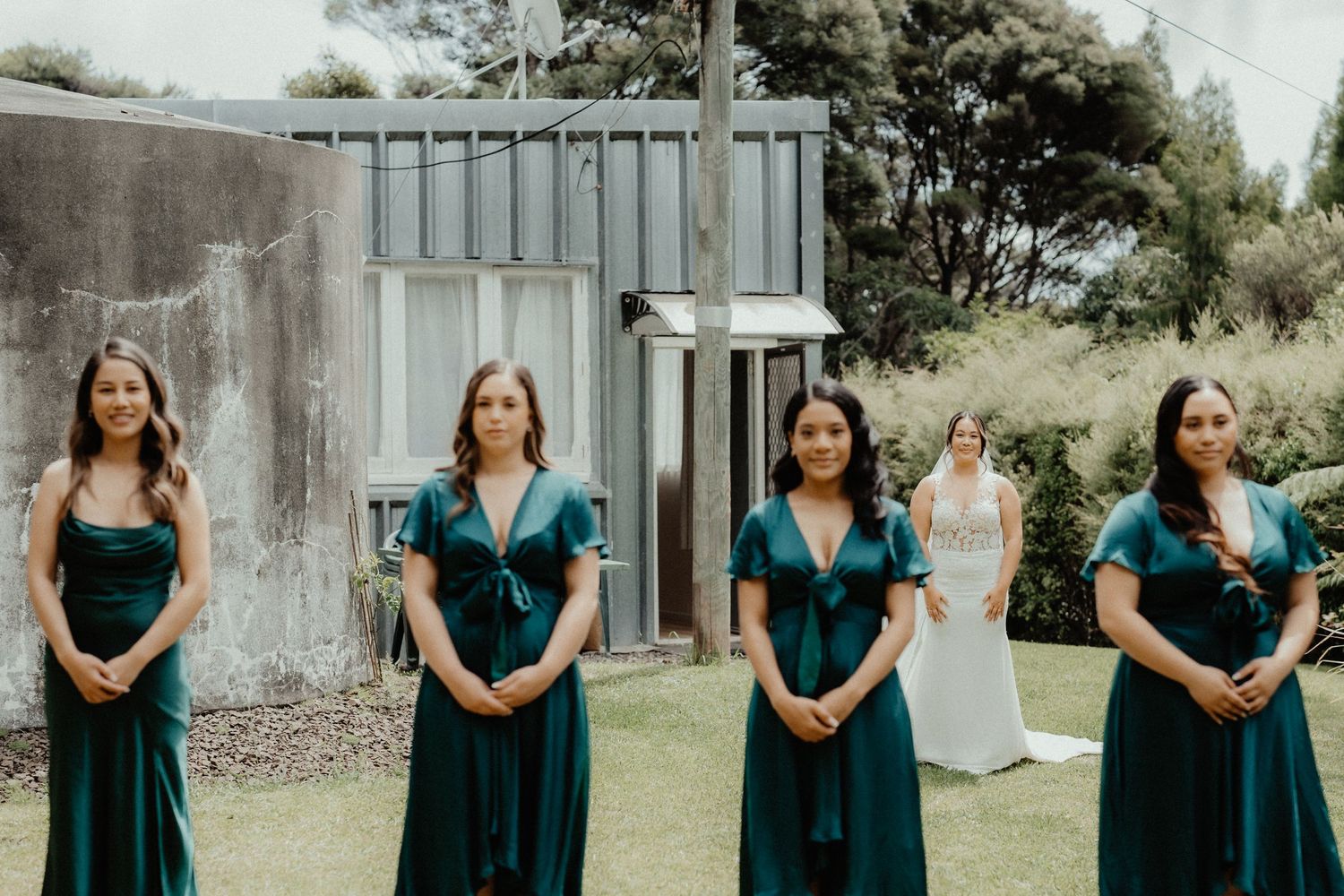 Four bridesmaids in emerald green dresses stand on the lawn while the bride in white waits behind them.