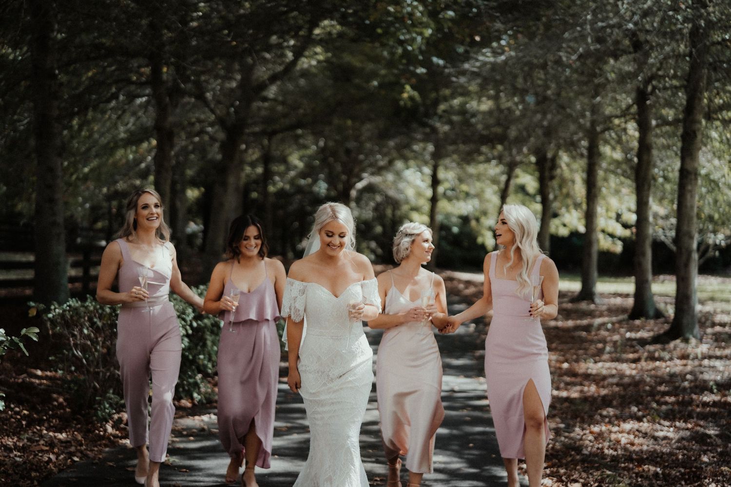Bride walks arm in arm with four smiling bridesmaids in blush dresses along a tree-lined path at a wedding.