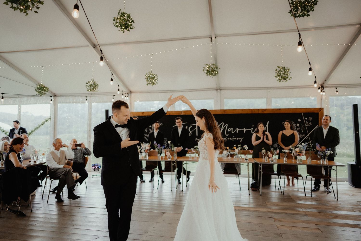 A bride and groom dance under a white marquee while wedding guests seated at long tables watch and celebrate.