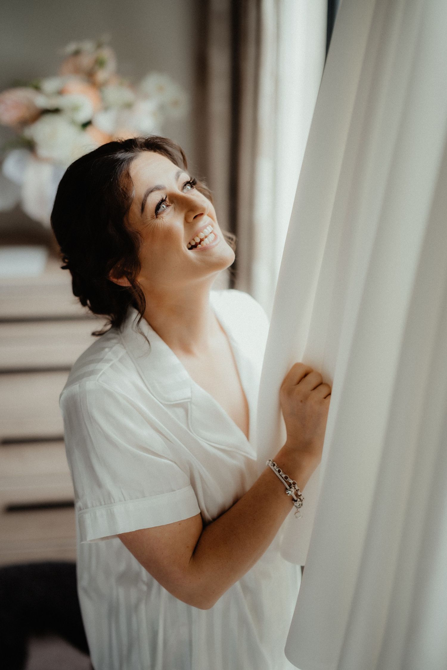 Smiling bride in a white satin robe gazes up at the light while holding a sheer curtain on her wedding morning.