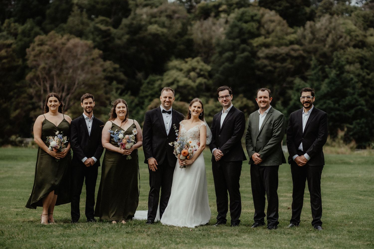 Bride, groom and wedding party in dark suits and green dresses pose in a line on a grassy lawn in front of trees.