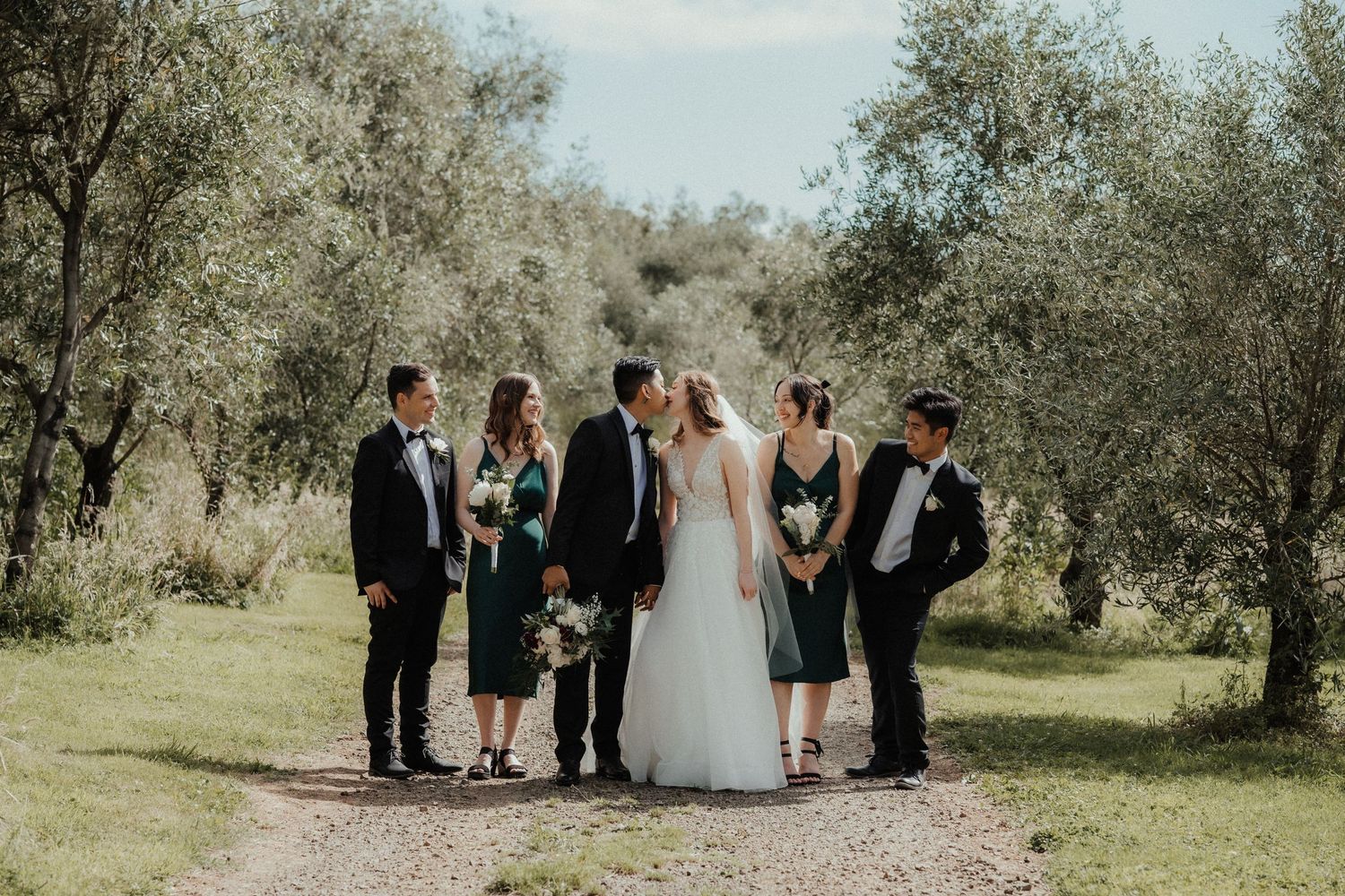 Bride and groom walk arm in arm along an olive grove path with bridesmaids and groomsmen following in formal wedding attire.