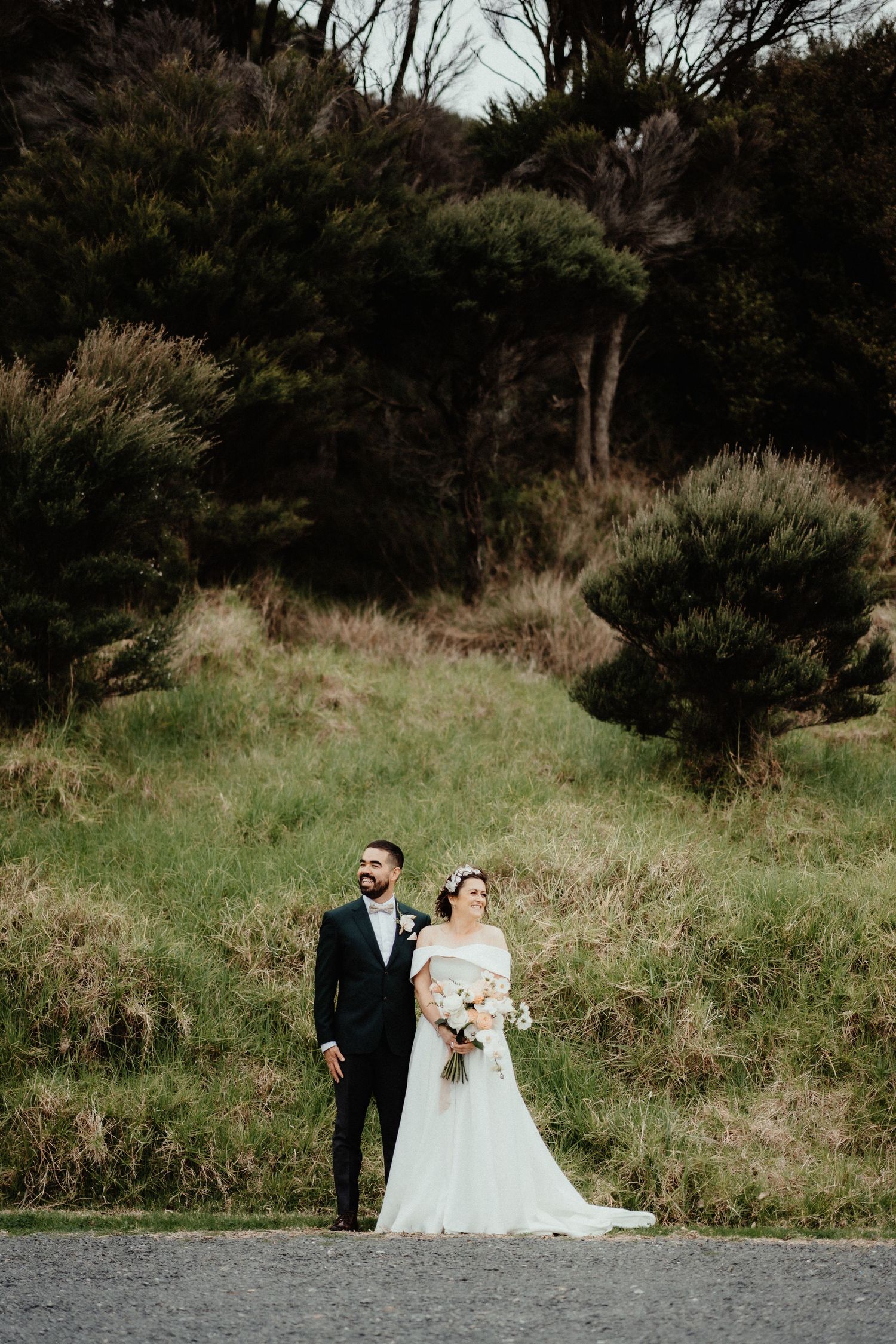 Bride holding a bouquet and groom in a dark suit stand together on a gravel path in front of a green grassy bank and trees.