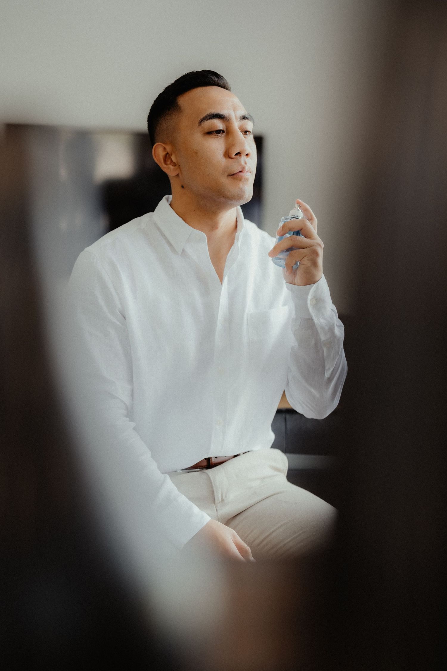 A groom in a white linen shirt sprays cologne while getting ready on his wedding morning.