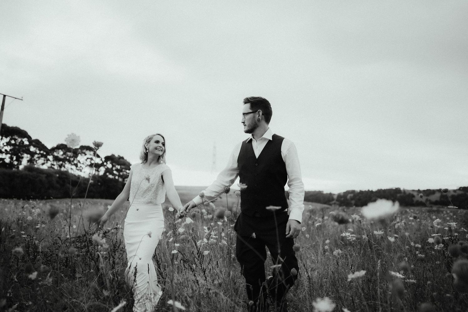 Black and white photo of a bride and groom holding hands and walking through a wildflower field on their wedding day.