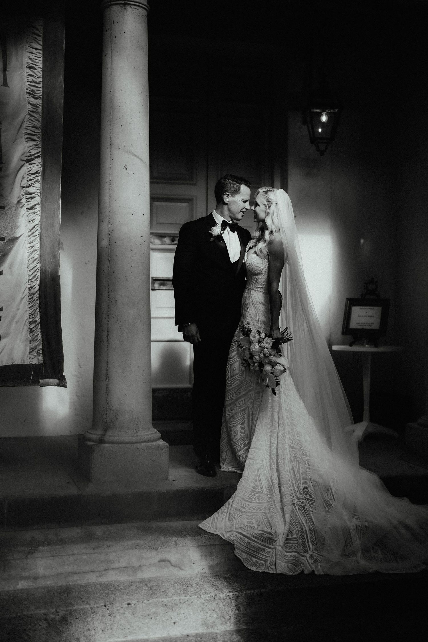 Black and white wedding portrait of a bride in a lace gown with long veil and a groom in a tuxedo smiling beside a stone column.