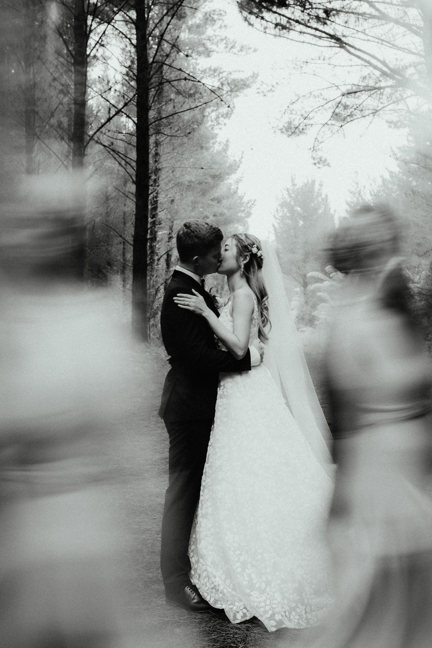 Bride and groom kiss on a forest path while blurred wedding guests move past in a black and white photograph.