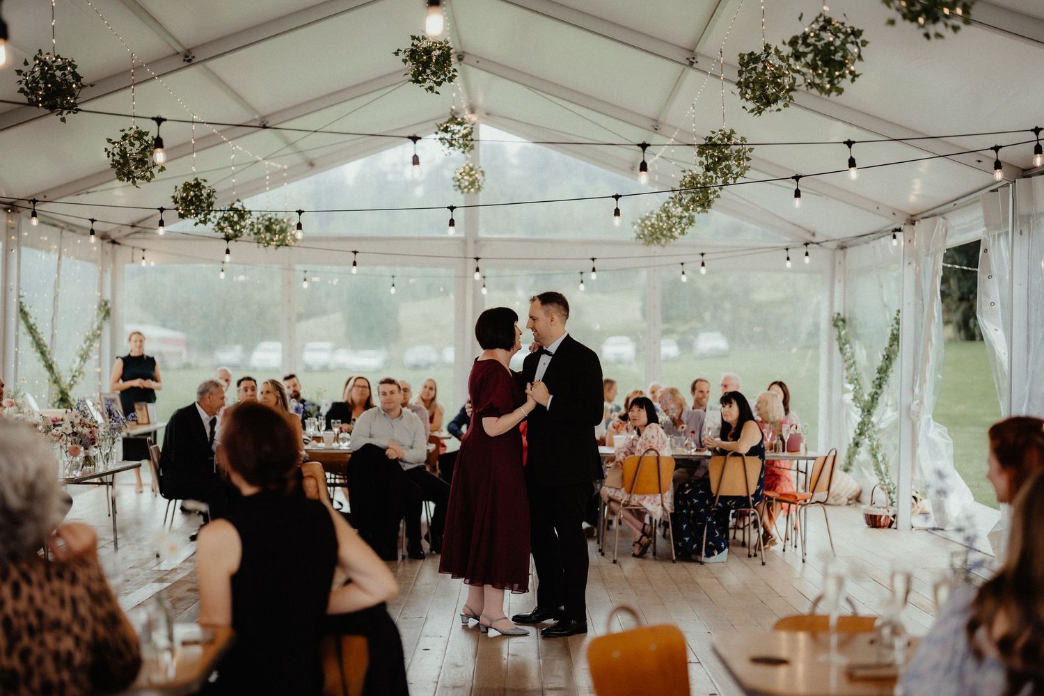 Groom in a black suit dances with a woman in a burgundy dress as seated guests watch inside a marquee decorated with greenery and lights.