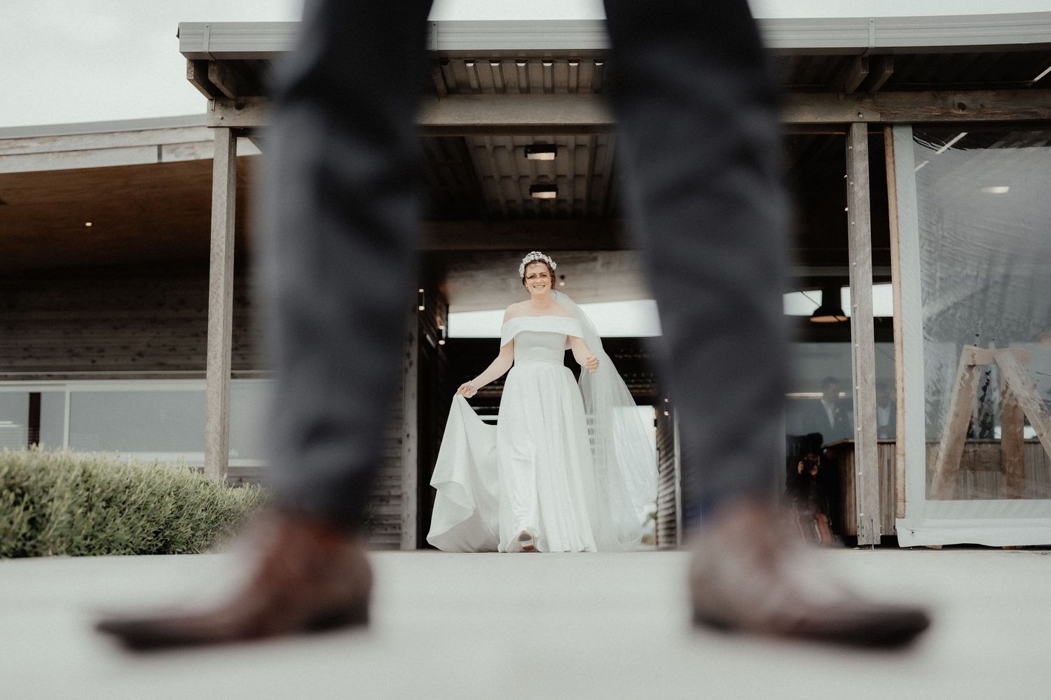 Bride in a long white gown holds her skirt in the distance, framed between the groom's legs and brown leather boots in the foreground.