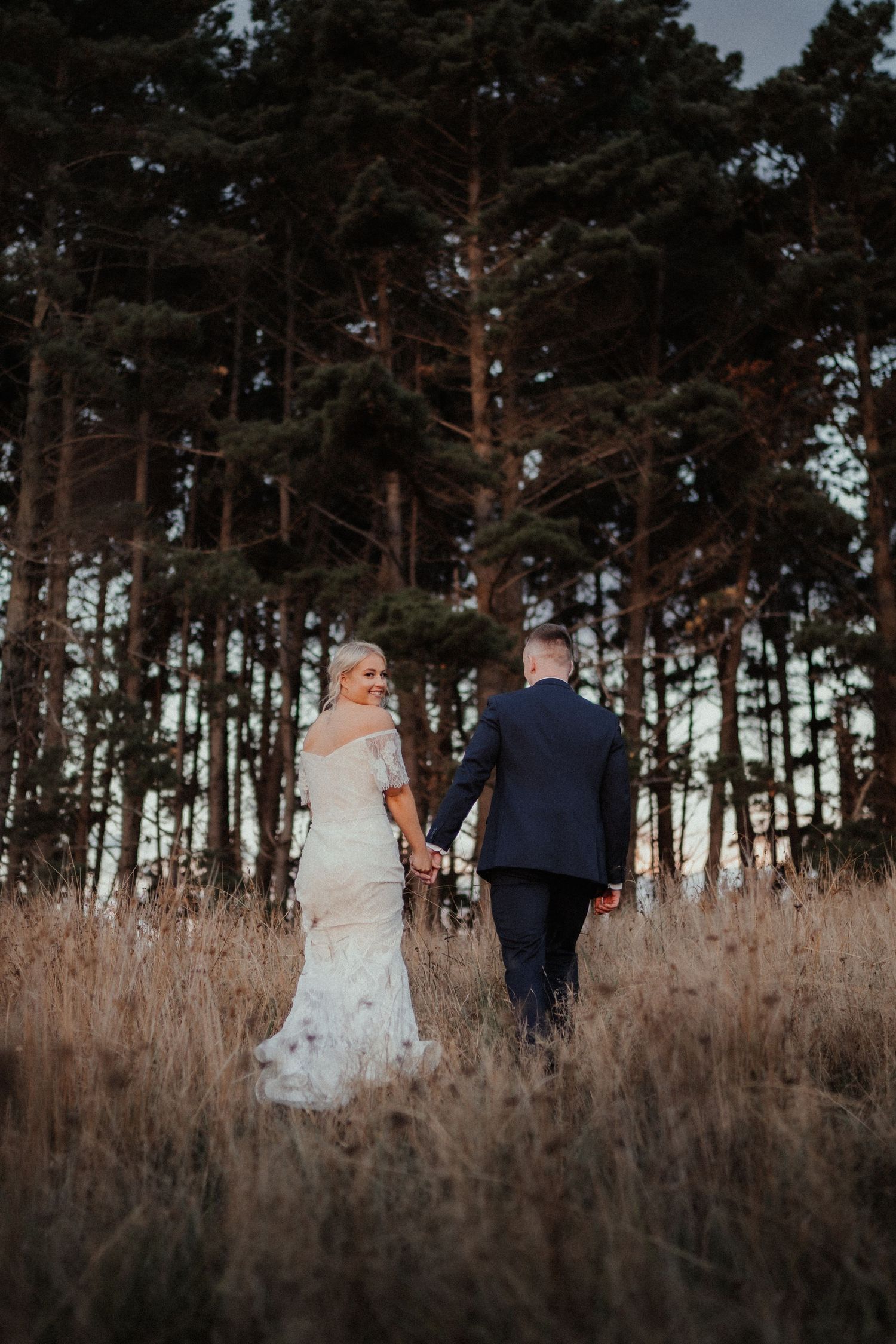 Bride in a lace wedding dress and groom in a navy suit hold hands and walk through tall dry grass toward a pine forest.