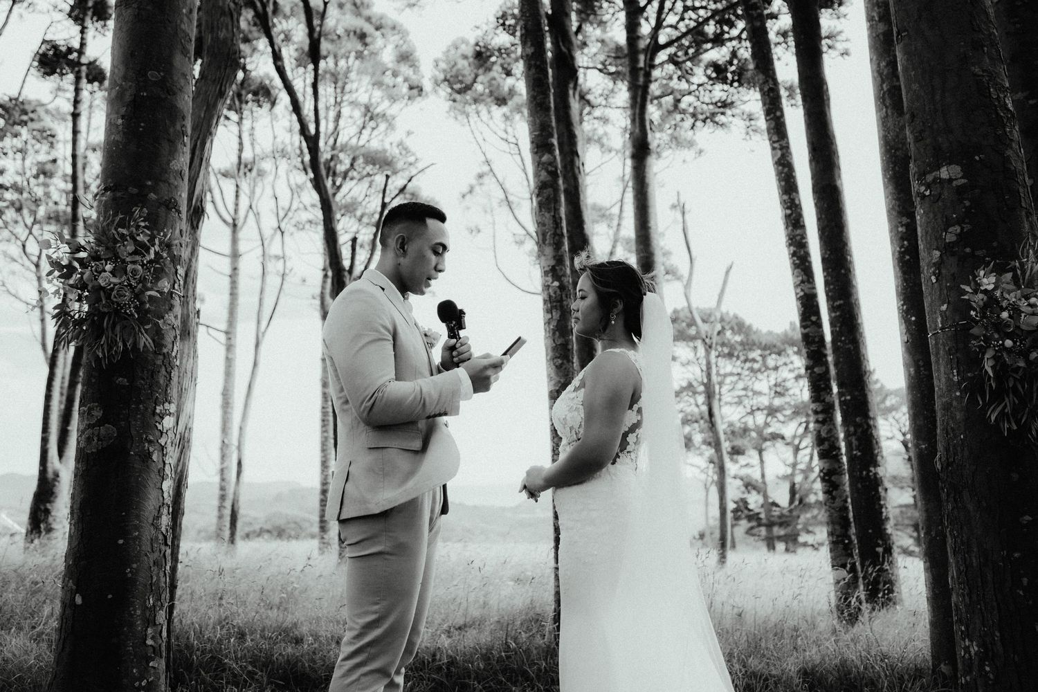 Groom reads vows into a microphone while facing the veiled bride among tall forest trees in a black and white wedding photo.