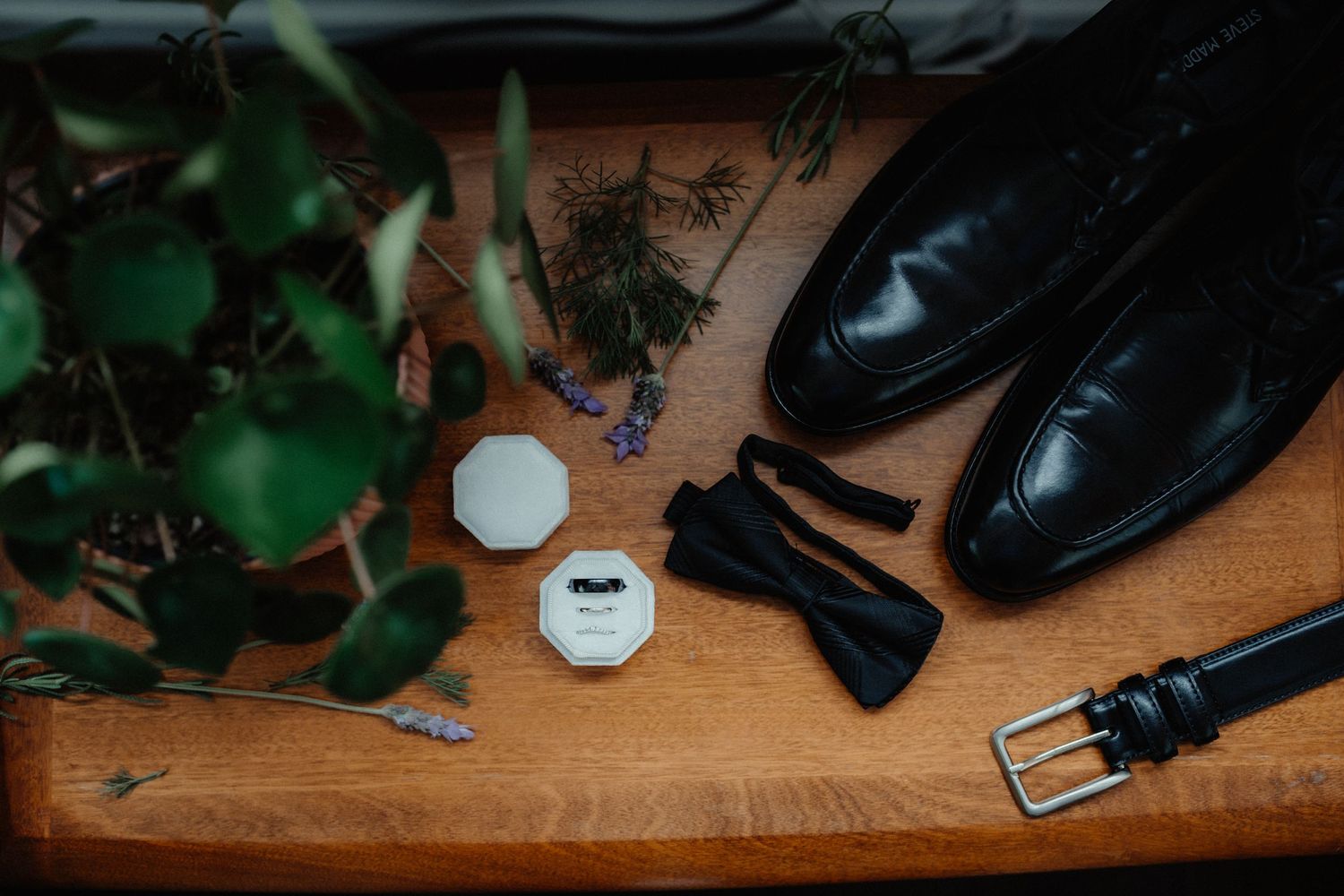 Black leather shoes, a bow tie, a belt, ring boxes and greenery sprigs arranged on a wooden surface for a wedding.