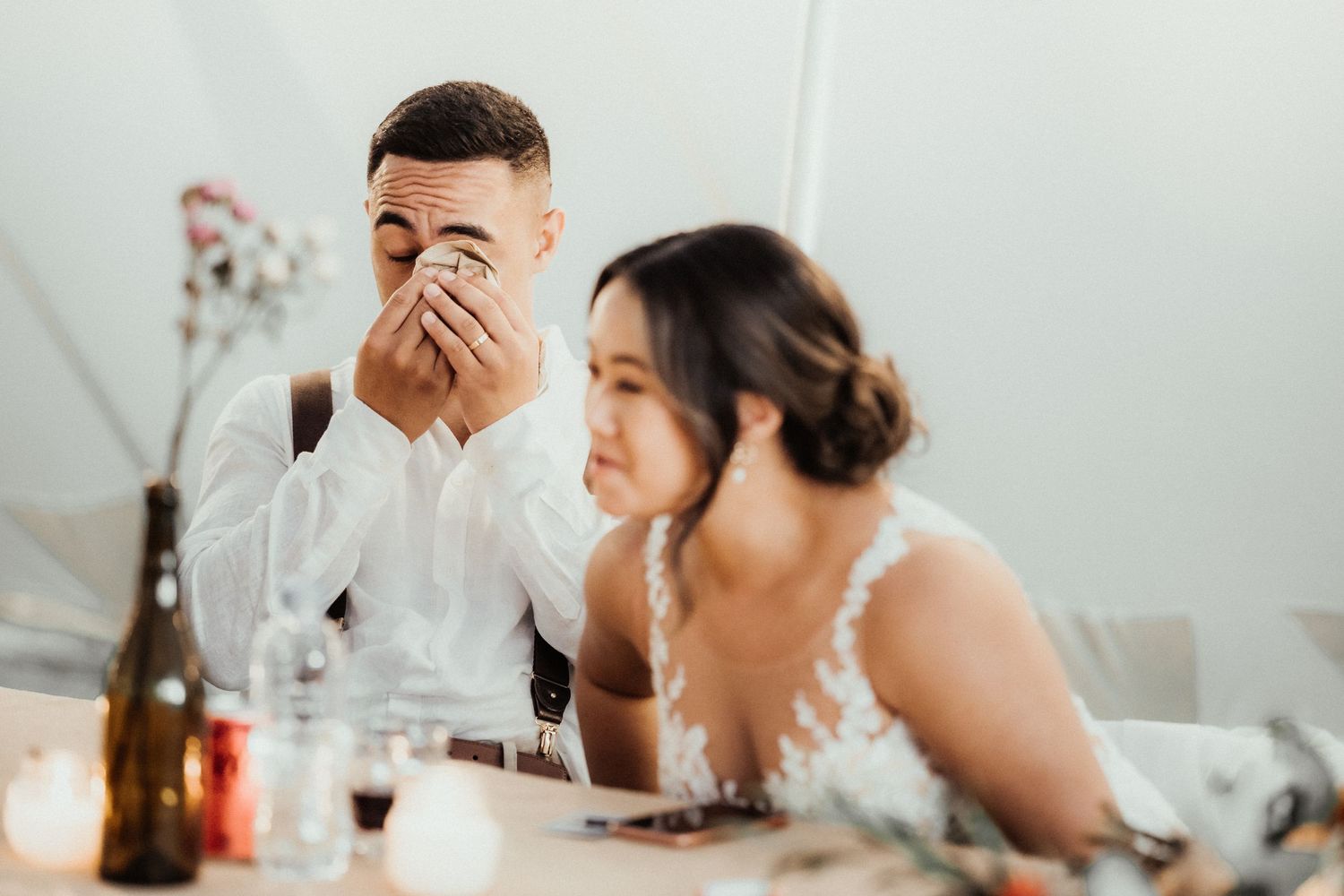 Emotional groom wipes his eyes while the bride sits beside him at the reception table during wedding speeches.