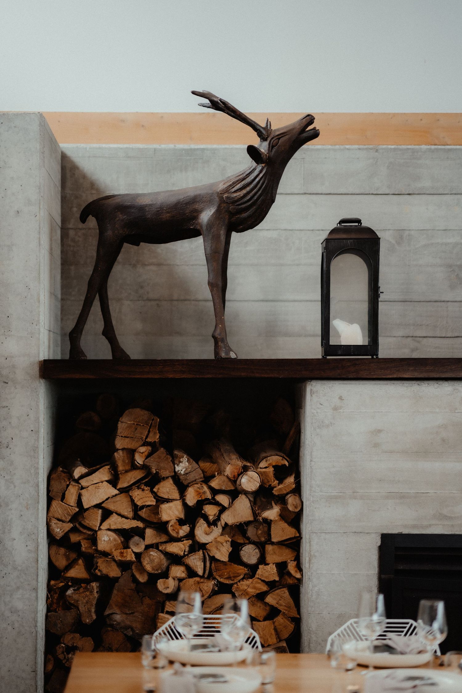 Rustic wooden deer sculpture and a black lantern sit above a neatly stacked firewood alcove in a modern concrete dining room.